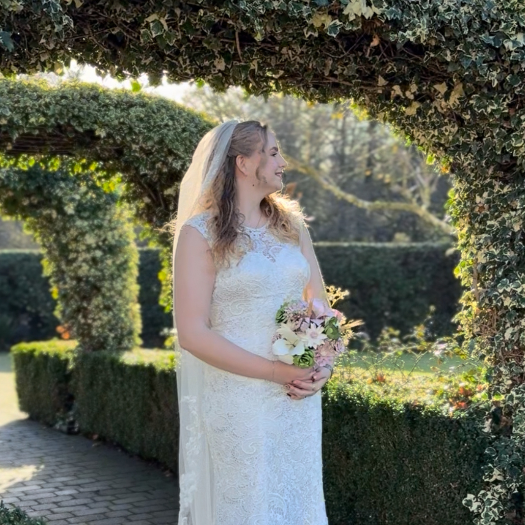 A bride in a white lace wedding dress holding a pink and white bouquet, standing in a garden under an archway covered with greenery, sunlight behind her.