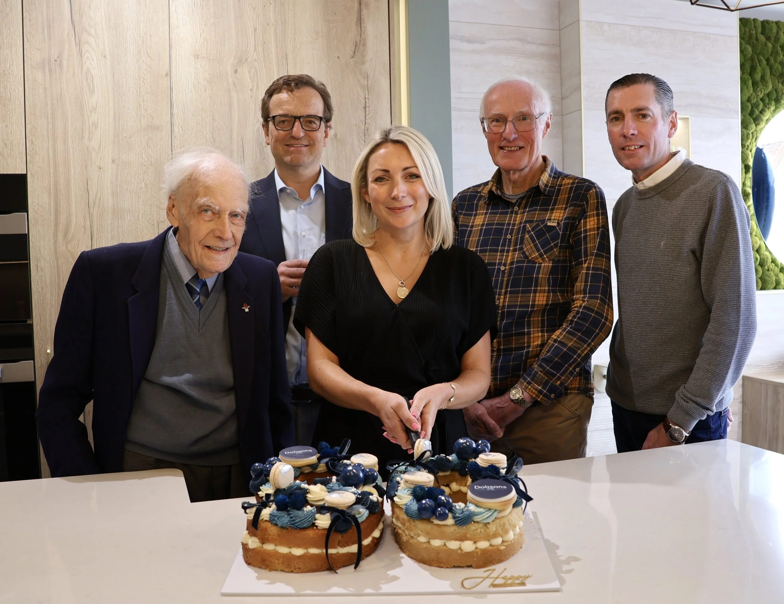 Six people standing behind a table with two decorated cakes, with a woman in black cutting into one of the cakes, in a modern indoor setting.