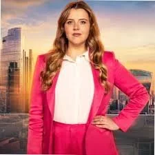 A woman with long wavy hair wearing a pink suit and white shirt standing outdoors with a cityscape and sunset in the background.