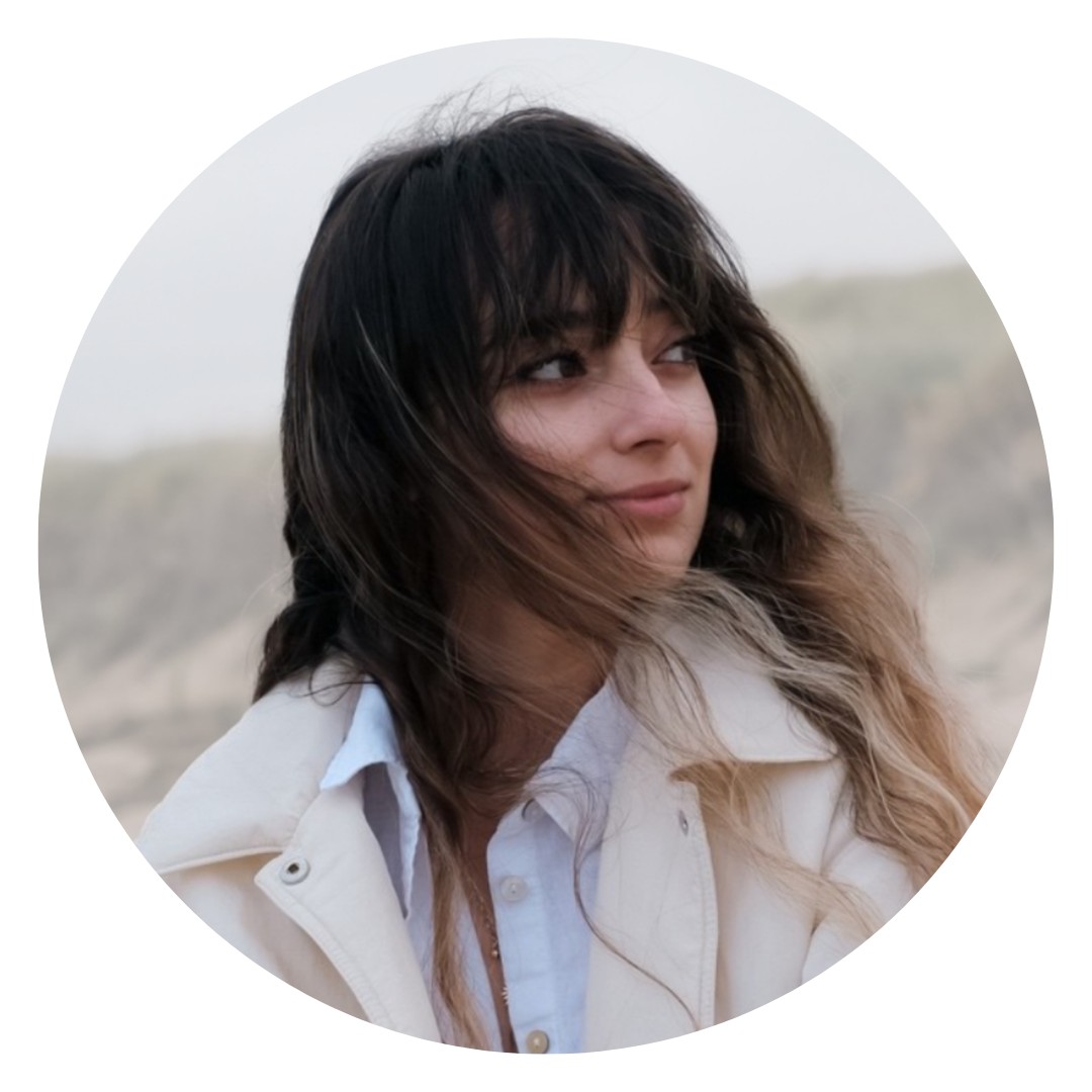 A woman with wavy, shoulder-length dark hair with light brown tips, wearing a white shirt and light-colored jacket, looking to the side, outdoors with a blurred background of dunes or rocky terrain.