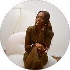 A woman in a brown outfit sitting on a white sofa with a white wall background.