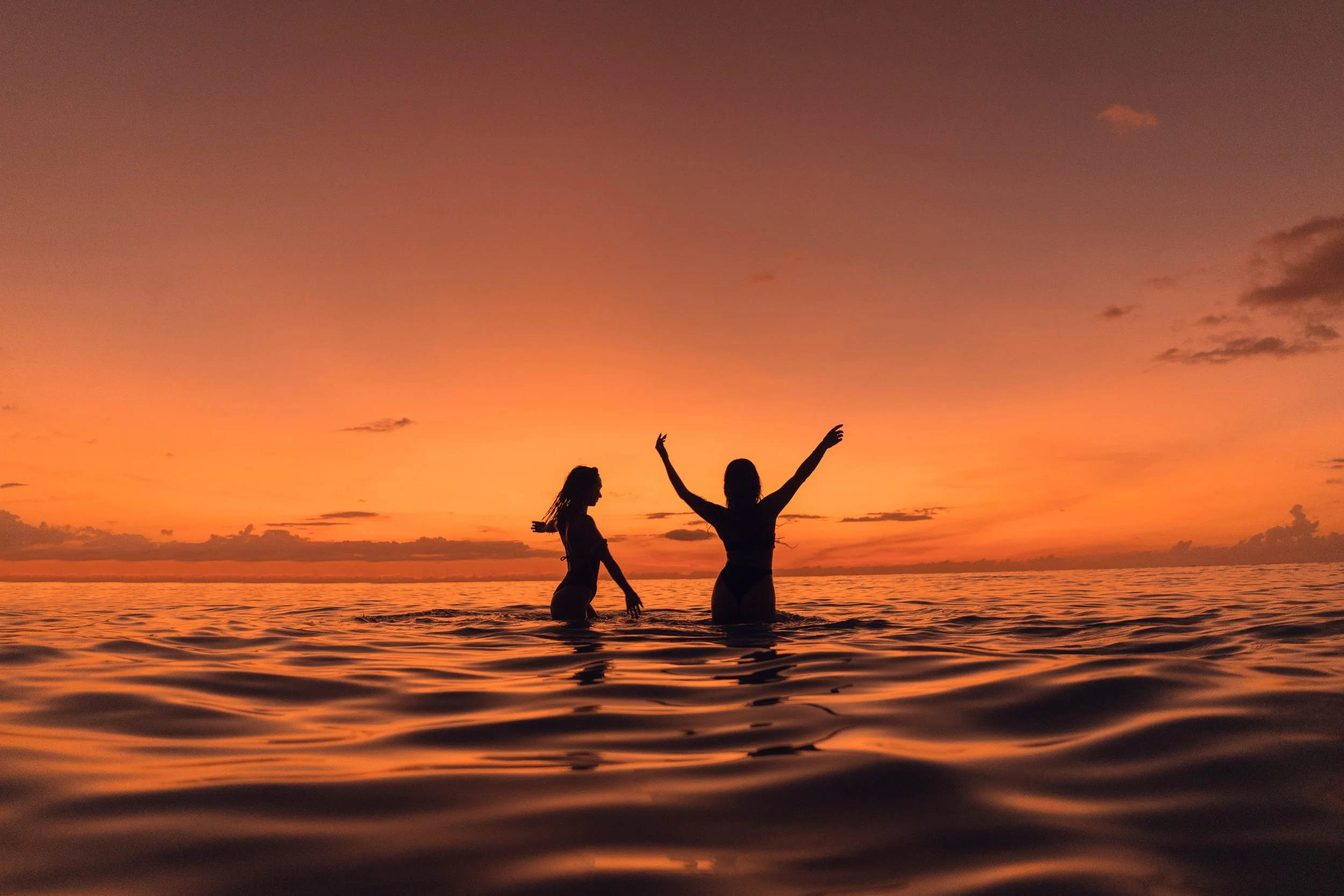 Two women in swimsuits standing in the water at sunset, one with arms raised, with a colorful sky and calm ocean in the background.