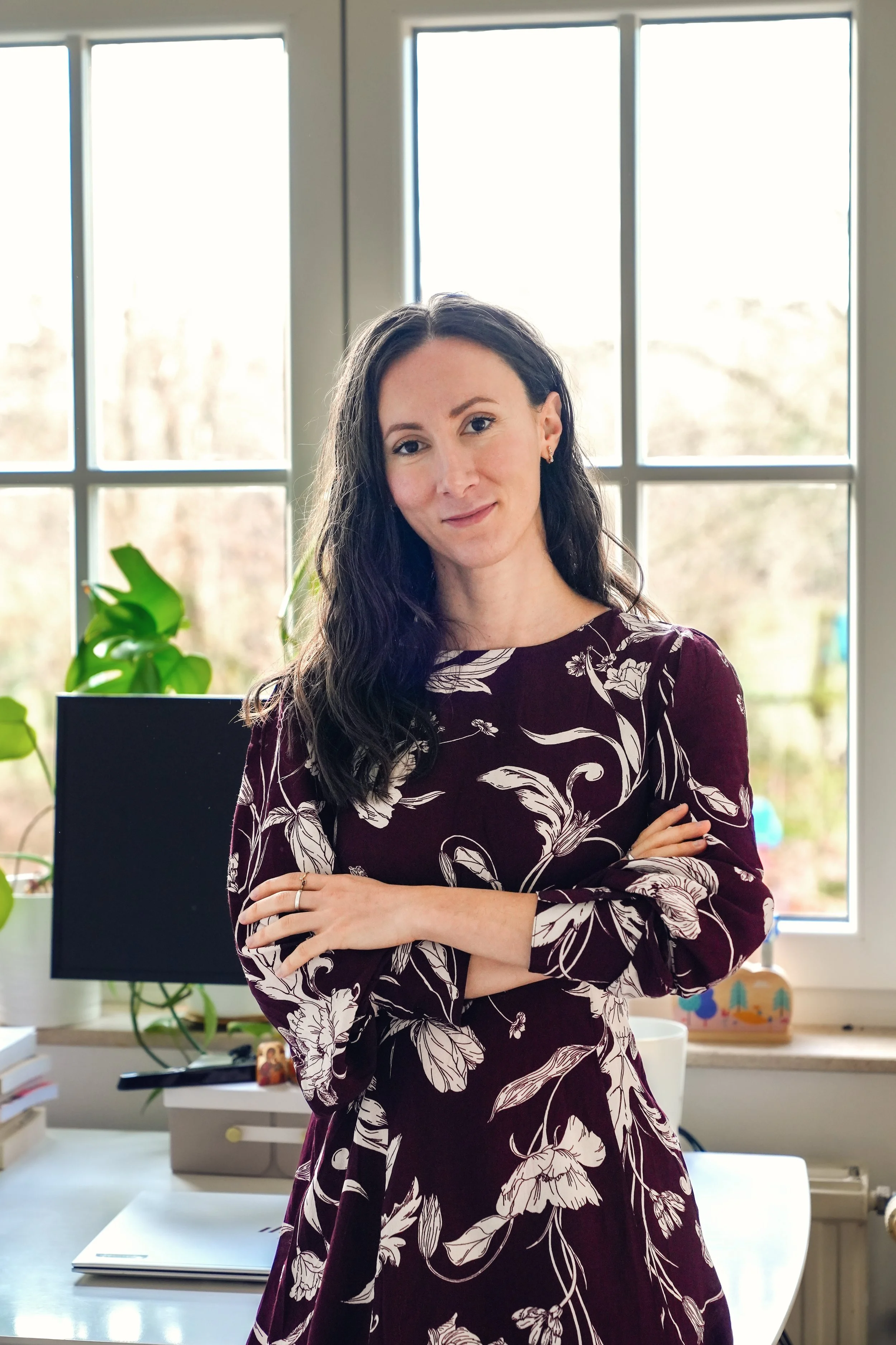 Femme avec cheveux longs et bouclés portant une robe à motifs floraux, debout dans un bureau devant une fenêtre, avec des plantes et un ordinateur en arrière-plan.
Mère, maman, post-partum, 4ème mois.