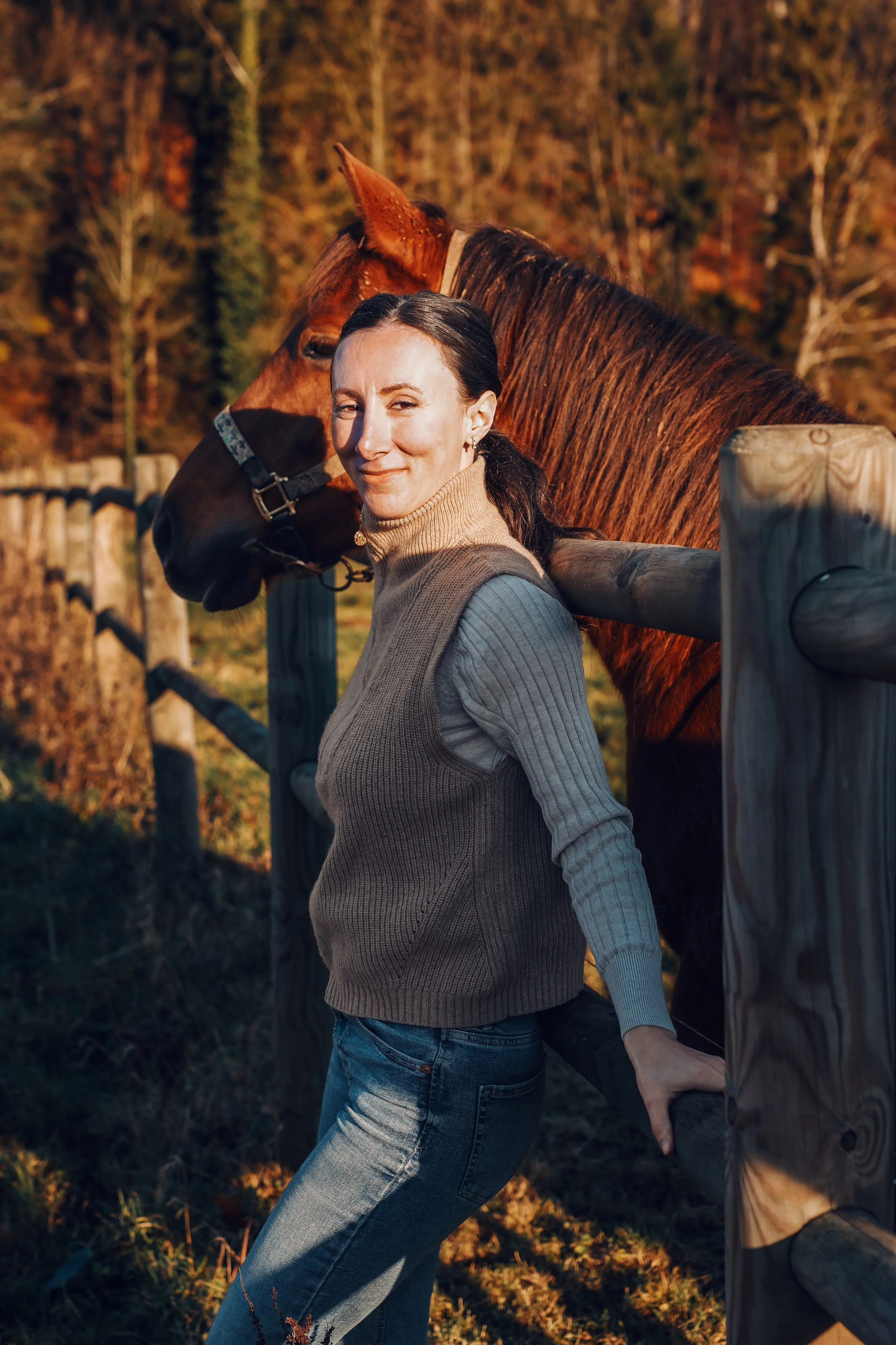 Une femme souriante en pull gris et jeans décontractés, posant à côté d'un cheval marron dans un champ en automne, avec des arbres en arrière-plan.
Mère, maman, post-partum, 4ème mois.