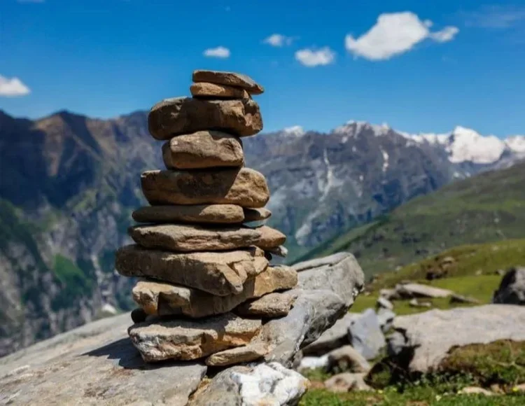 A scenic mountain landscape with two stone cairns in the foreground, green grass, rocks, small trees, and a mountain in the background.