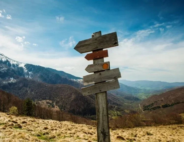 A group of eight men standing on rocky terrain at a summit, celebrating with hats raised in the air. There is a British flag on a pole nearby. The photo is black-and-white, with a hazy, cloud-filled sky in the background.