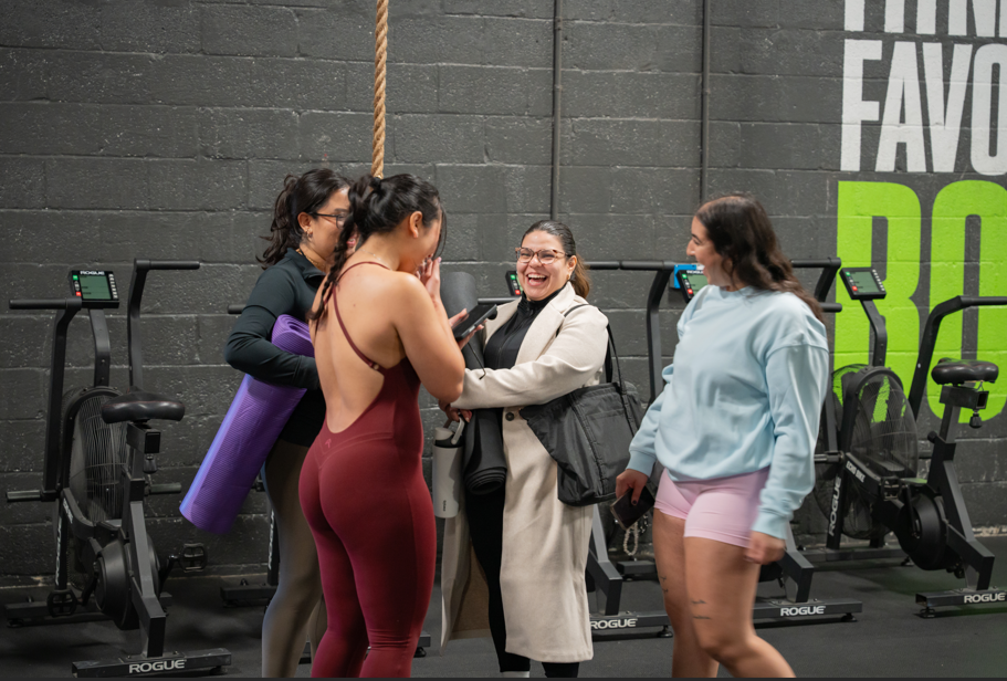 Group of four women in a gym, one in a yoga outfit, another reading her phone, three smiling and laughing together.