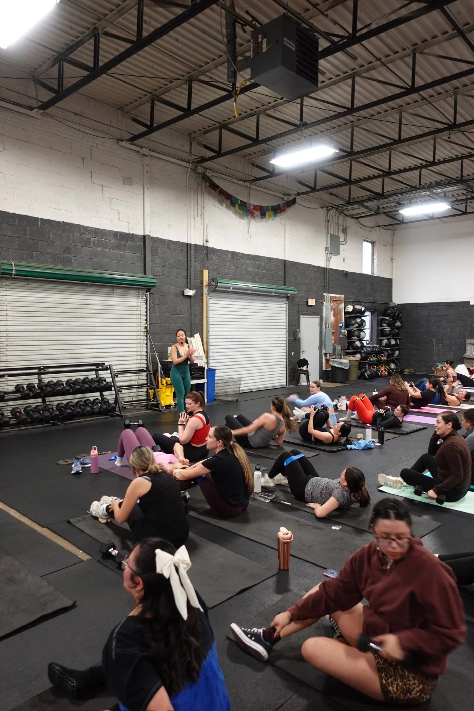 A group fitness class in Northern Virginia with women on exercise mats doing abdominal exercises in a gym with black walls, storage racks, and a high ceiling.