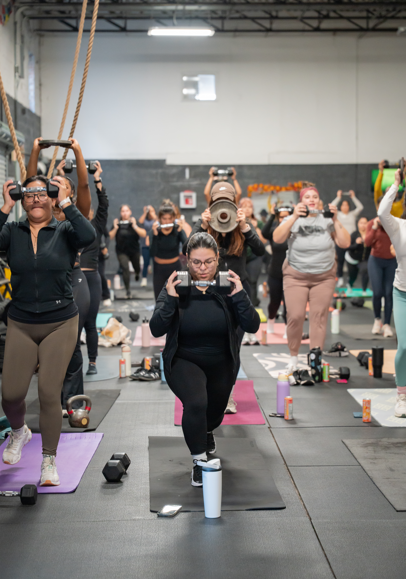 Group of women participating in a fitness class at a gym, with some holding kettlebells and others using other exercise equipment.