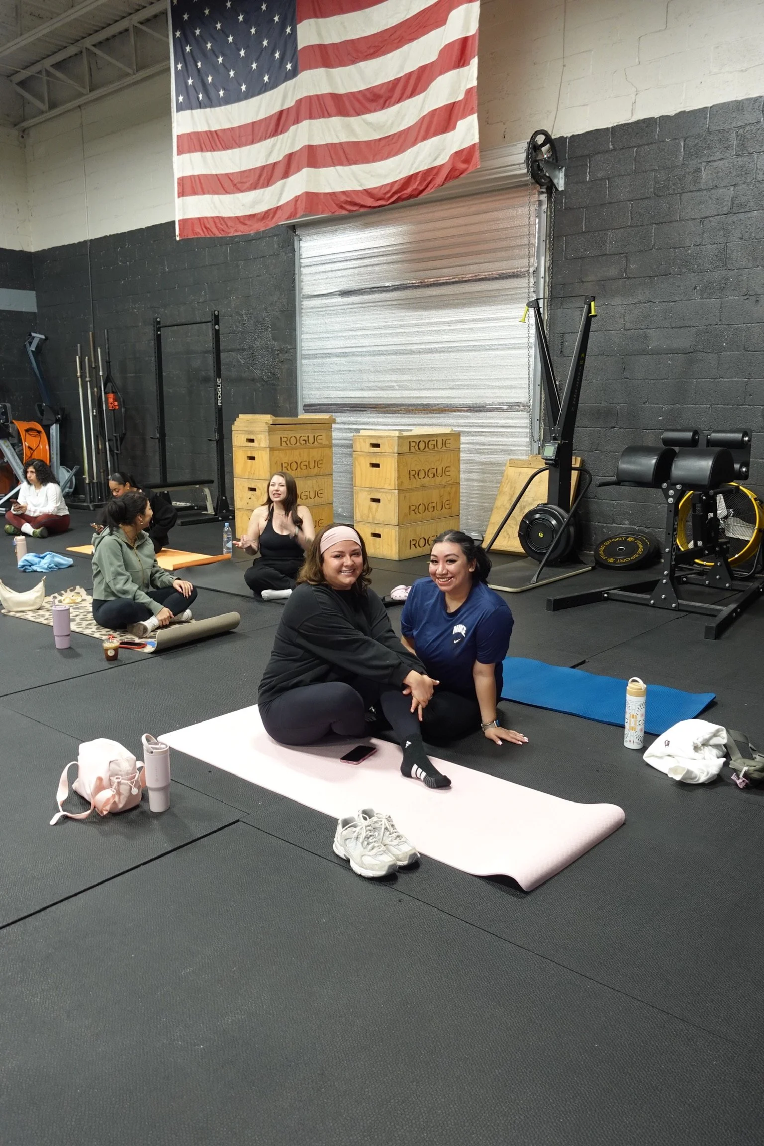 Group of women practicing yoga in a gym with black walls, an American flag, and gym equipment in the background.