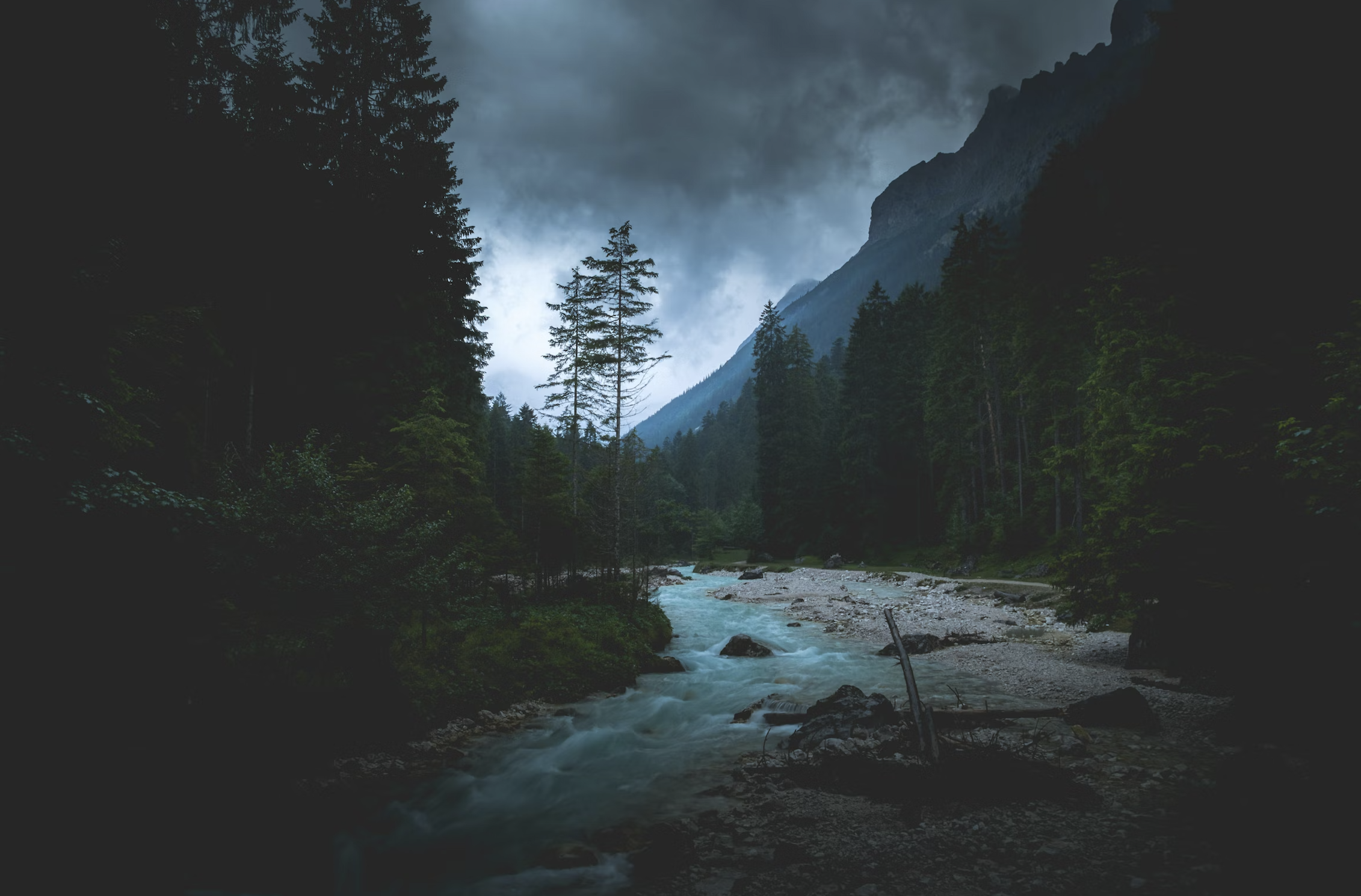 A river flowing through a forest with mountains and dark clouds in the background.