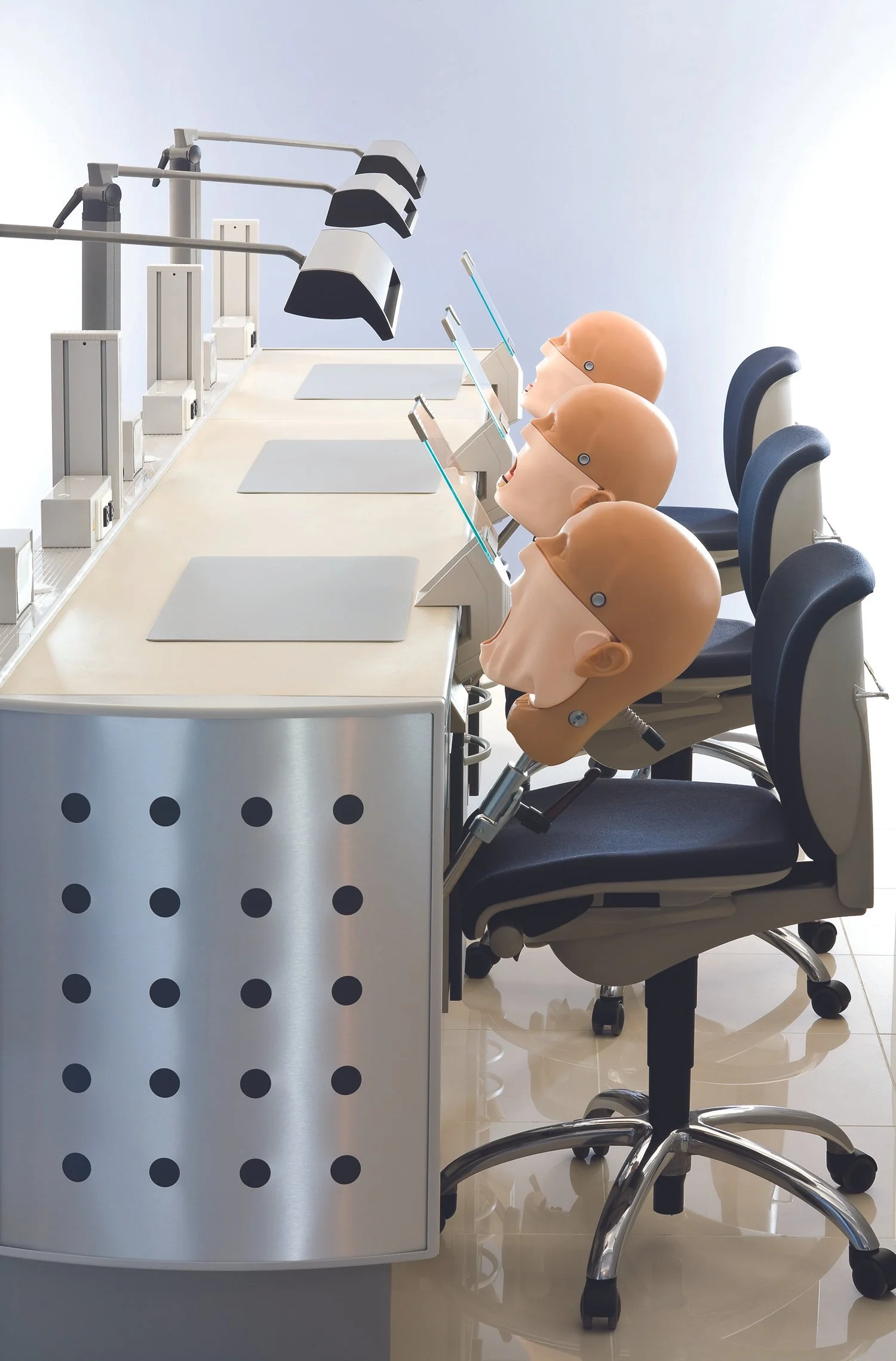 Three medical training mannequins with open mouths are seated at a row of chairs in front of a medical training station with simulated laryngoscopes and monitors.