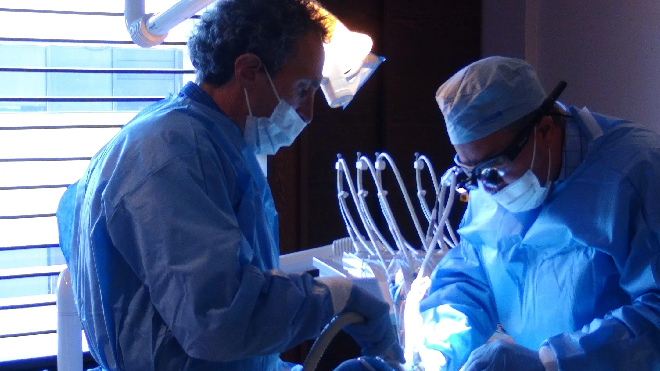 Two surgeons in blue surgical gowns, masks, and glasses perform a procedure in an operating room with focused lighting and medical equipment.