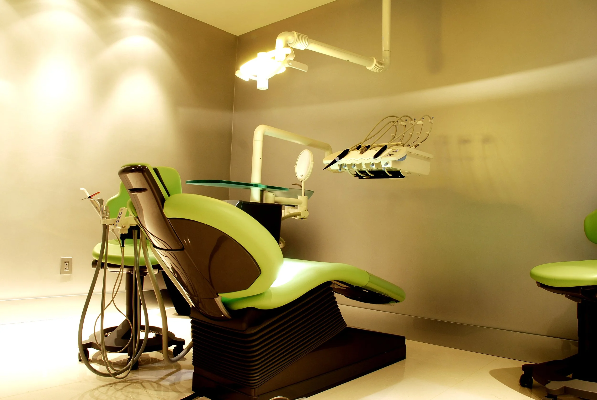 A modern dental examination room featuring a green dental chair with attached equipment, including dental tools, a light, and a small table, with beige walls and flooring.