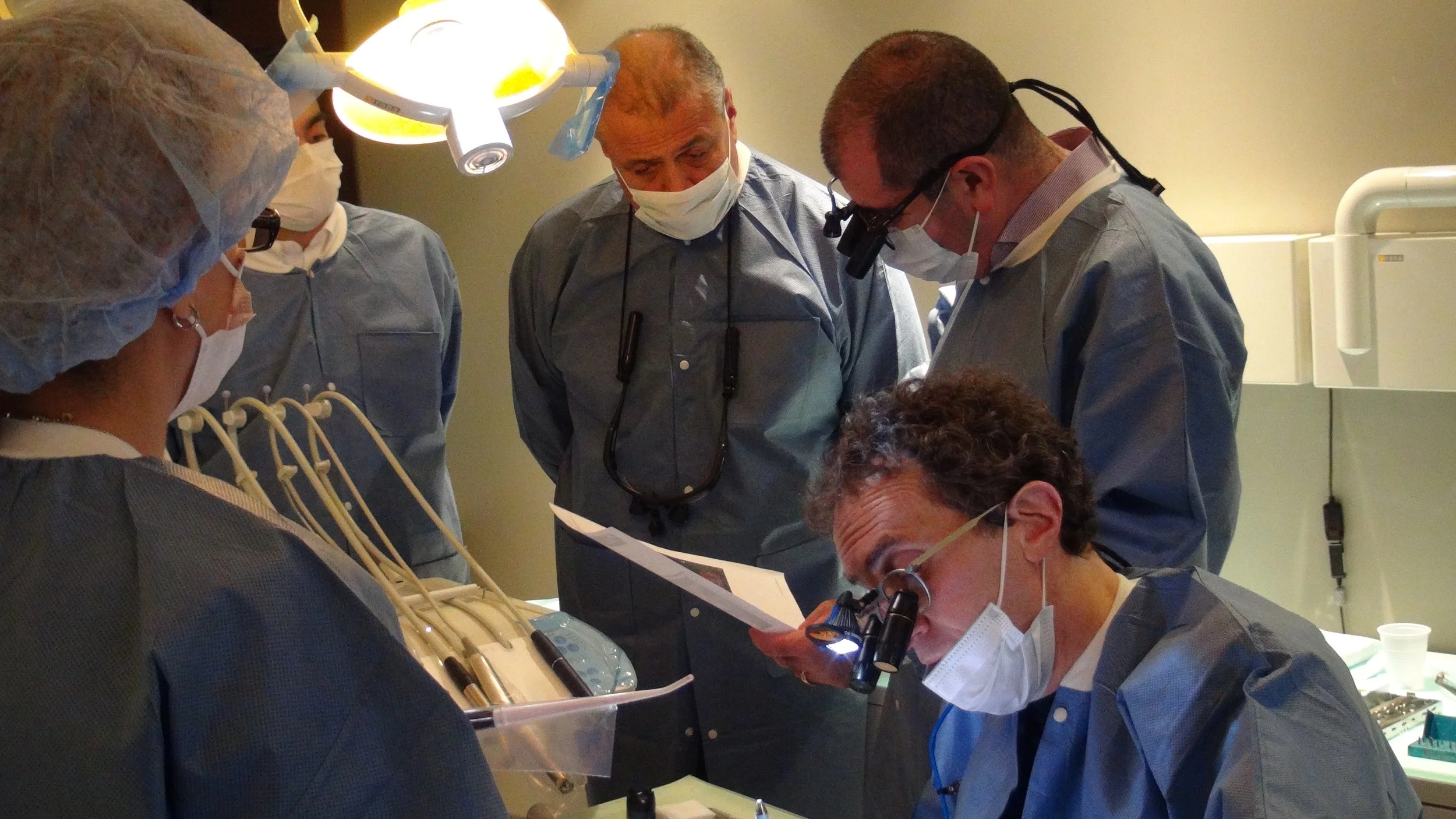 Group of medical professionals performing a surgical procedure in an operating room, wearing scrubs, masks, and protective eyewear, with surgical lights overhead.