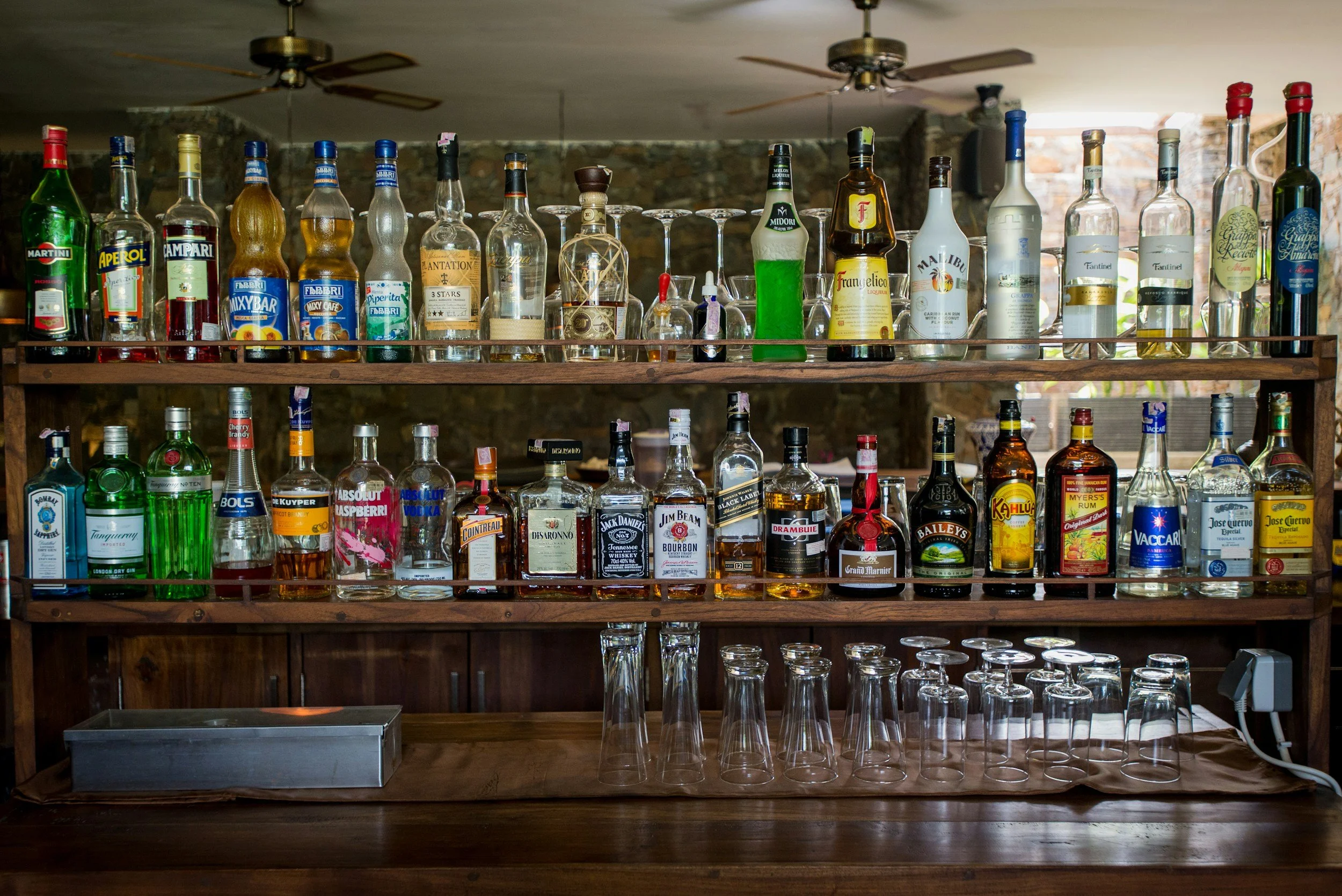 A wooden bar shelf with various bottles of liquor on top and glasses below, against a background of a brick wall and two ceiling fans.