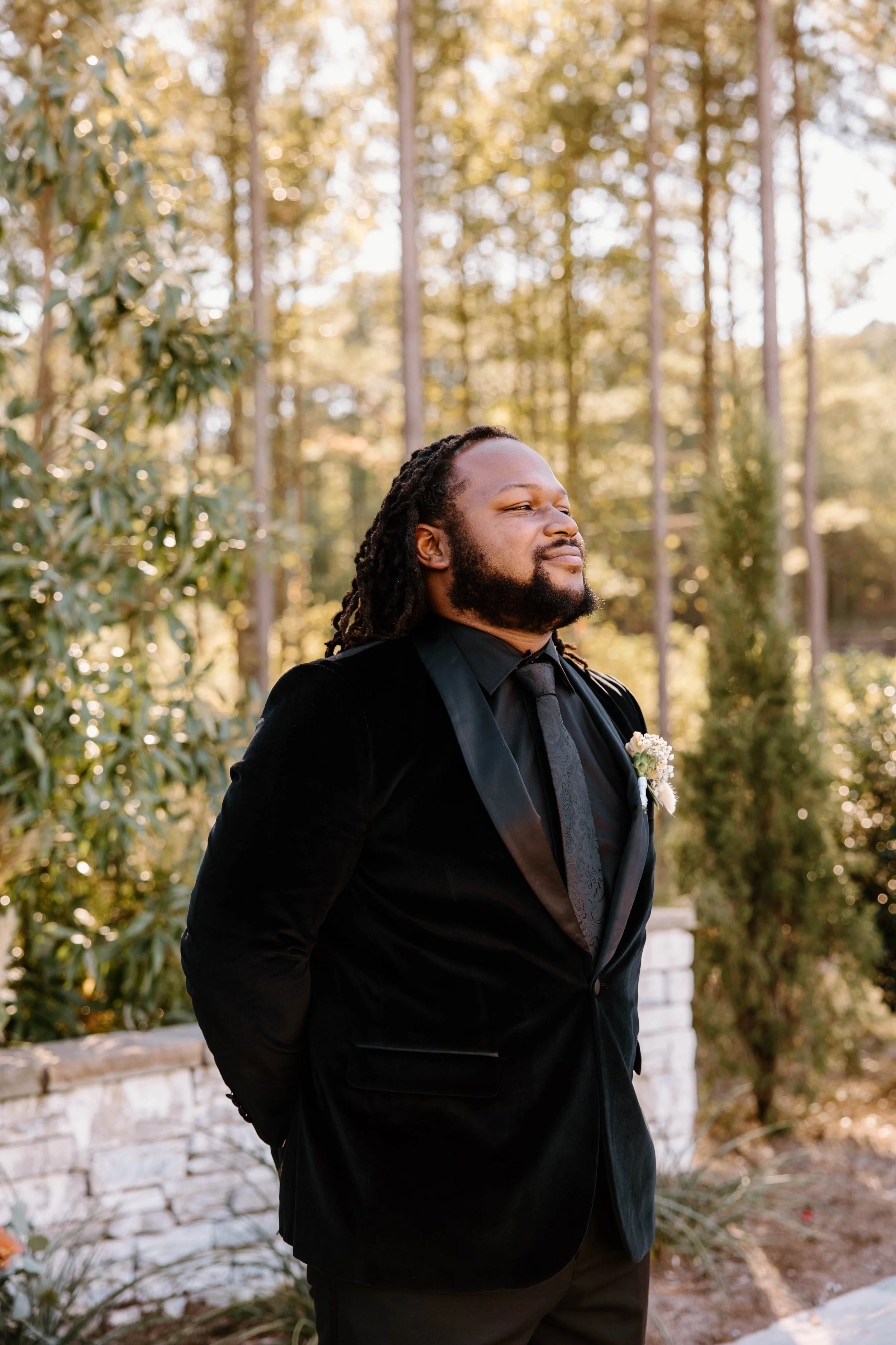 A man in a black tuxedo with a boutonnière standing outdoors among trees, with his eyes closed and a slight smile.