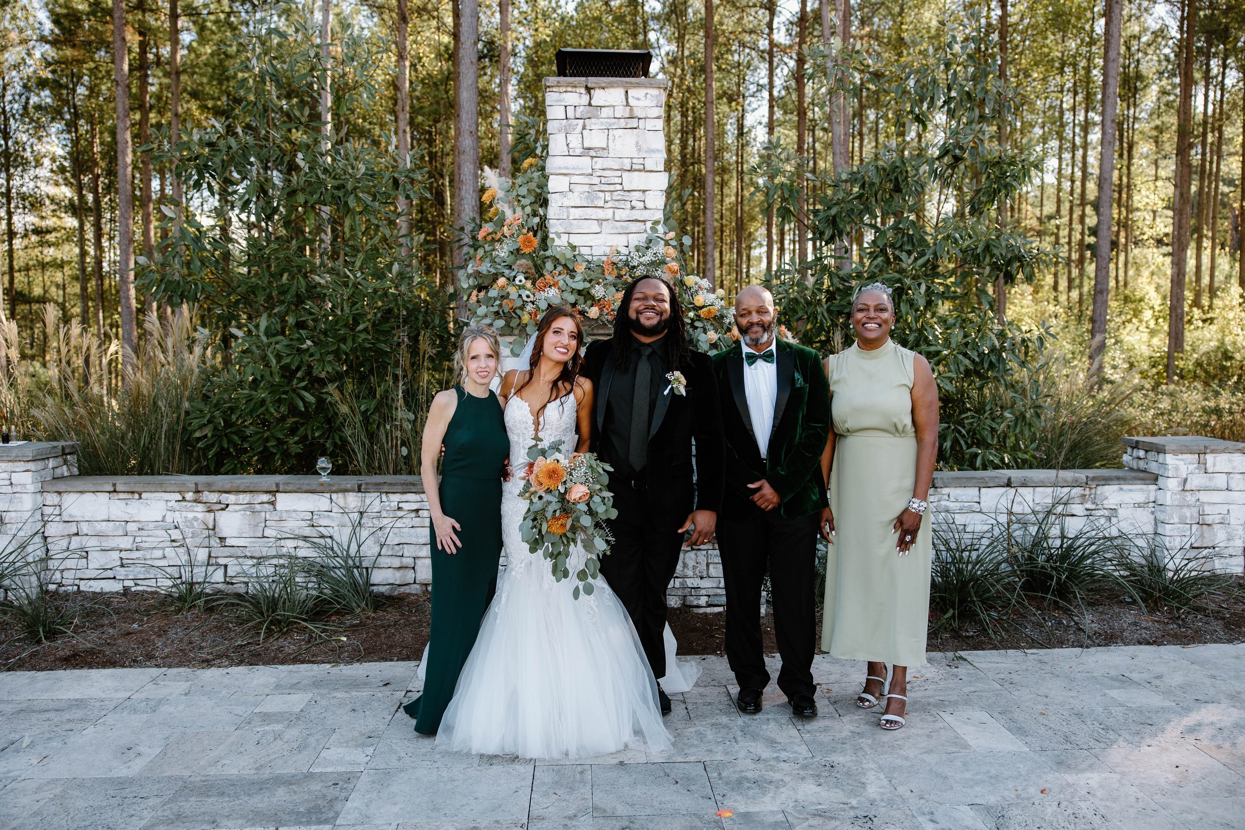 Group of five people at a wedding, standing outdoors in front of a stone fireplace with flowers, trees, and greenery.