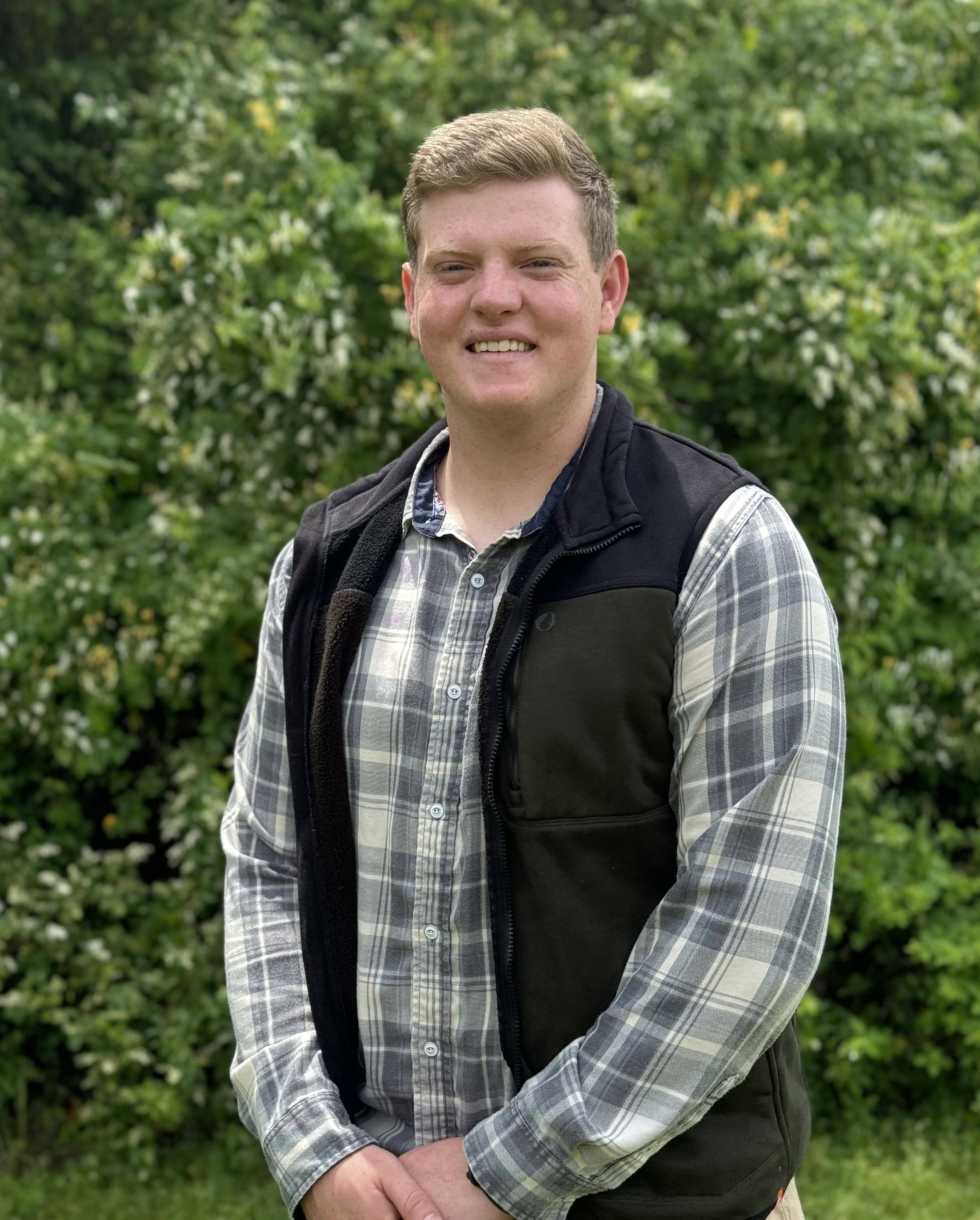 Drew Montgomery - Seek General Contractors - Young man standing outdoors in front of green bushes, smiling, wearing a plaid shirt and black vest.