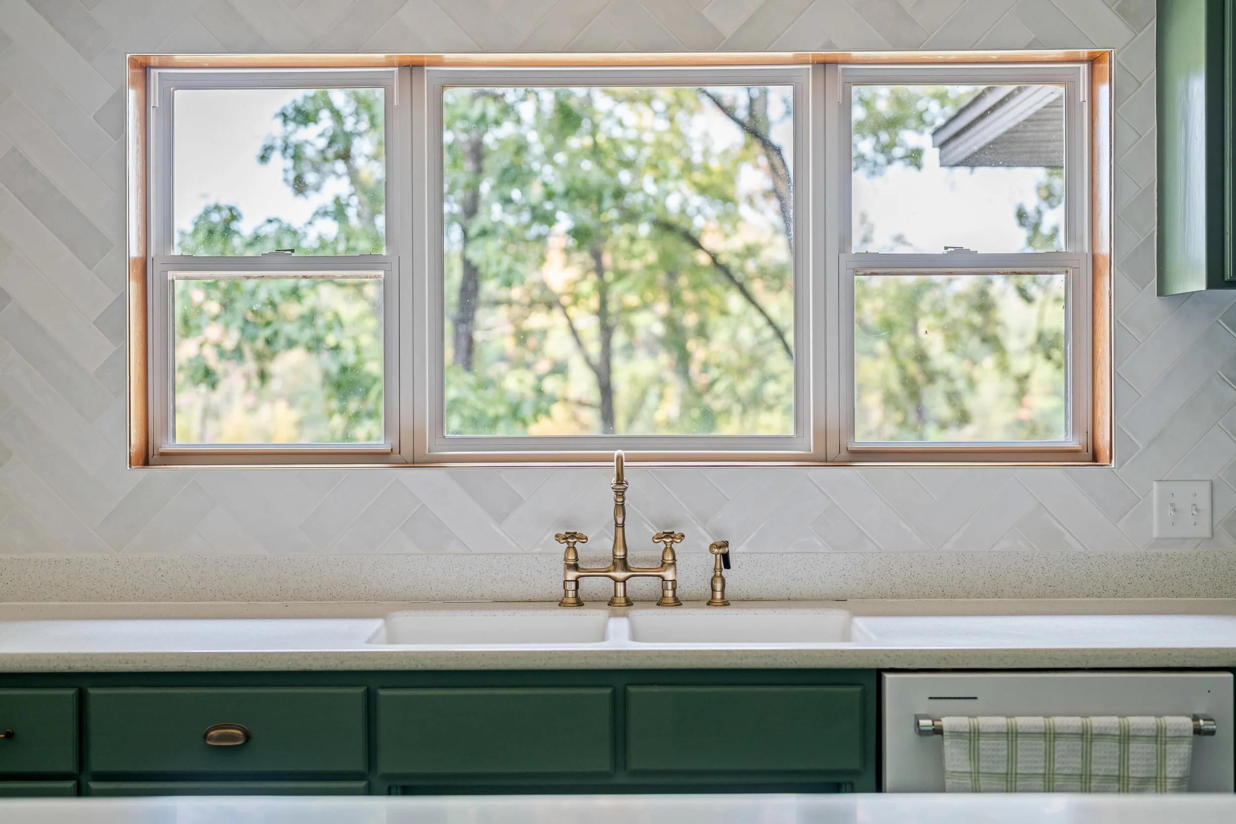 Kitchen window above a sink, with a view of green trees outside. Green cabinets and a white countertop with a brass faucet and fixtures.