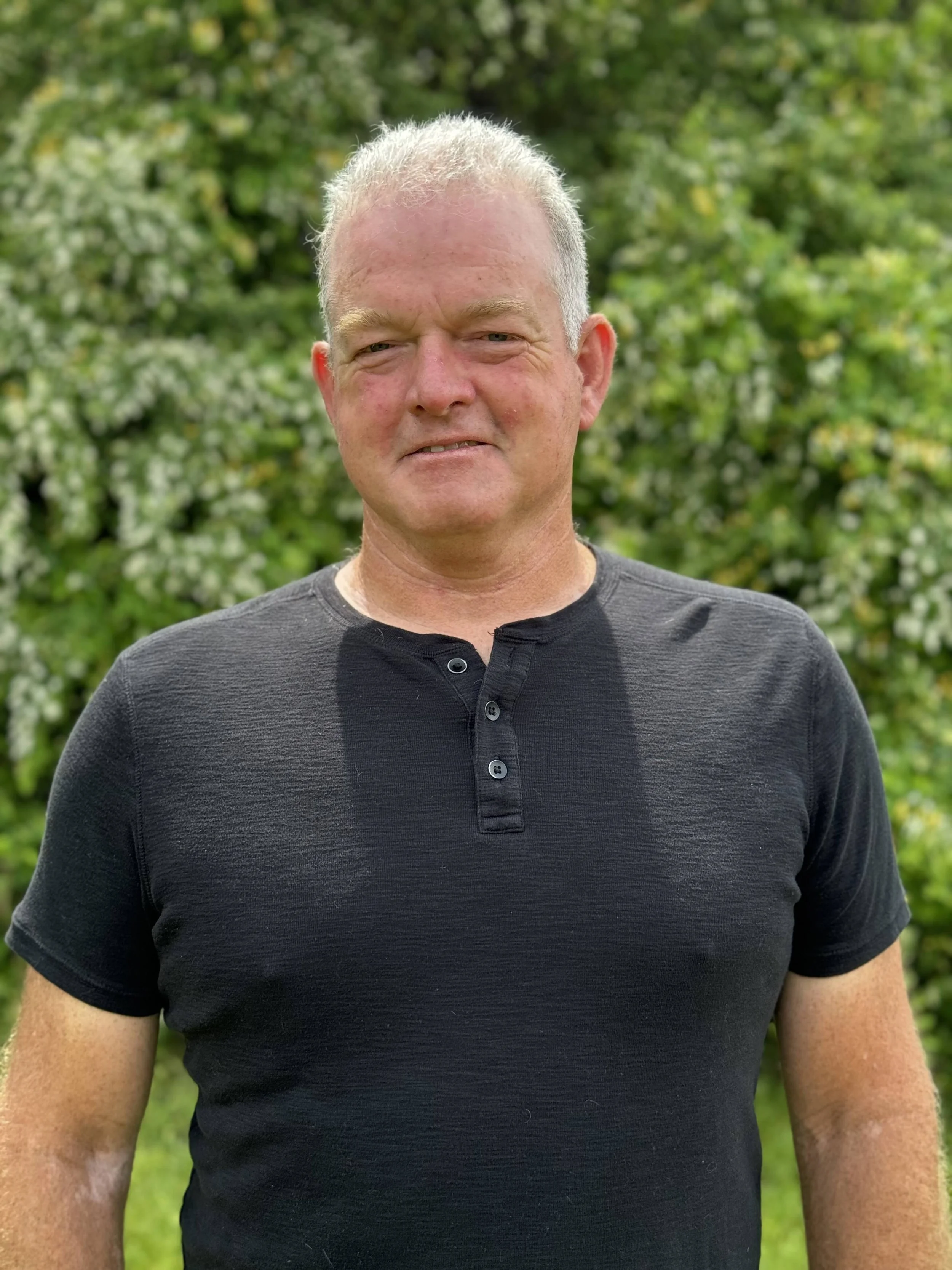 Jack Montgomery - Seek General Contractors - Man standing outdoors in front of green trees wearing a black short-sleeve shirt.