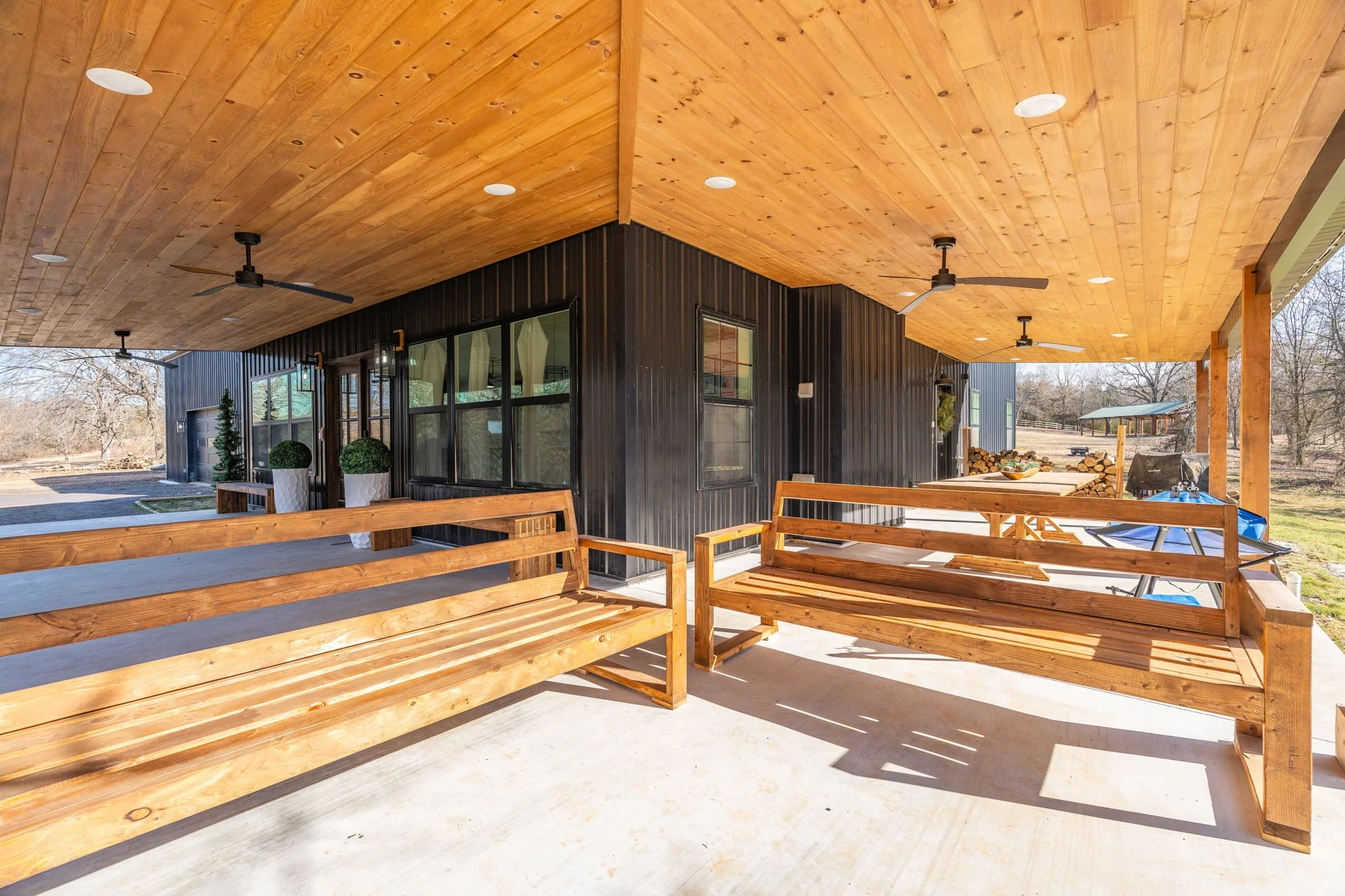 Covered patio with wooden benches, a ceiling fan, and outdoor chairs and tables, attached to a modern house with black exterior walls and large windows, with trees and open land in the background.