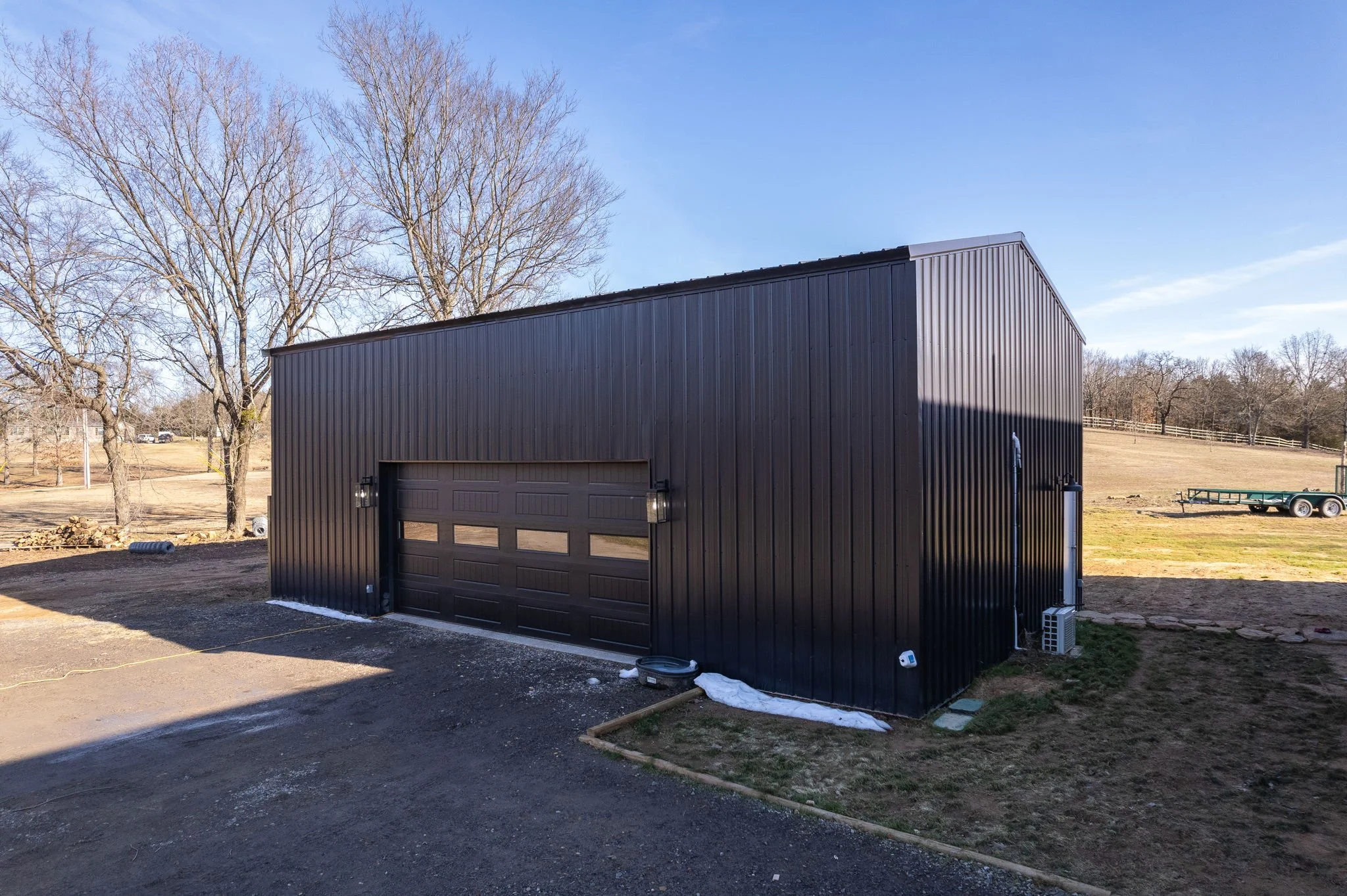 A black metal garage or shed with a closed sectional roll-up door, situated outdoors on a patch of dirt and grass, with leafless trees and a blue sky in the background.
