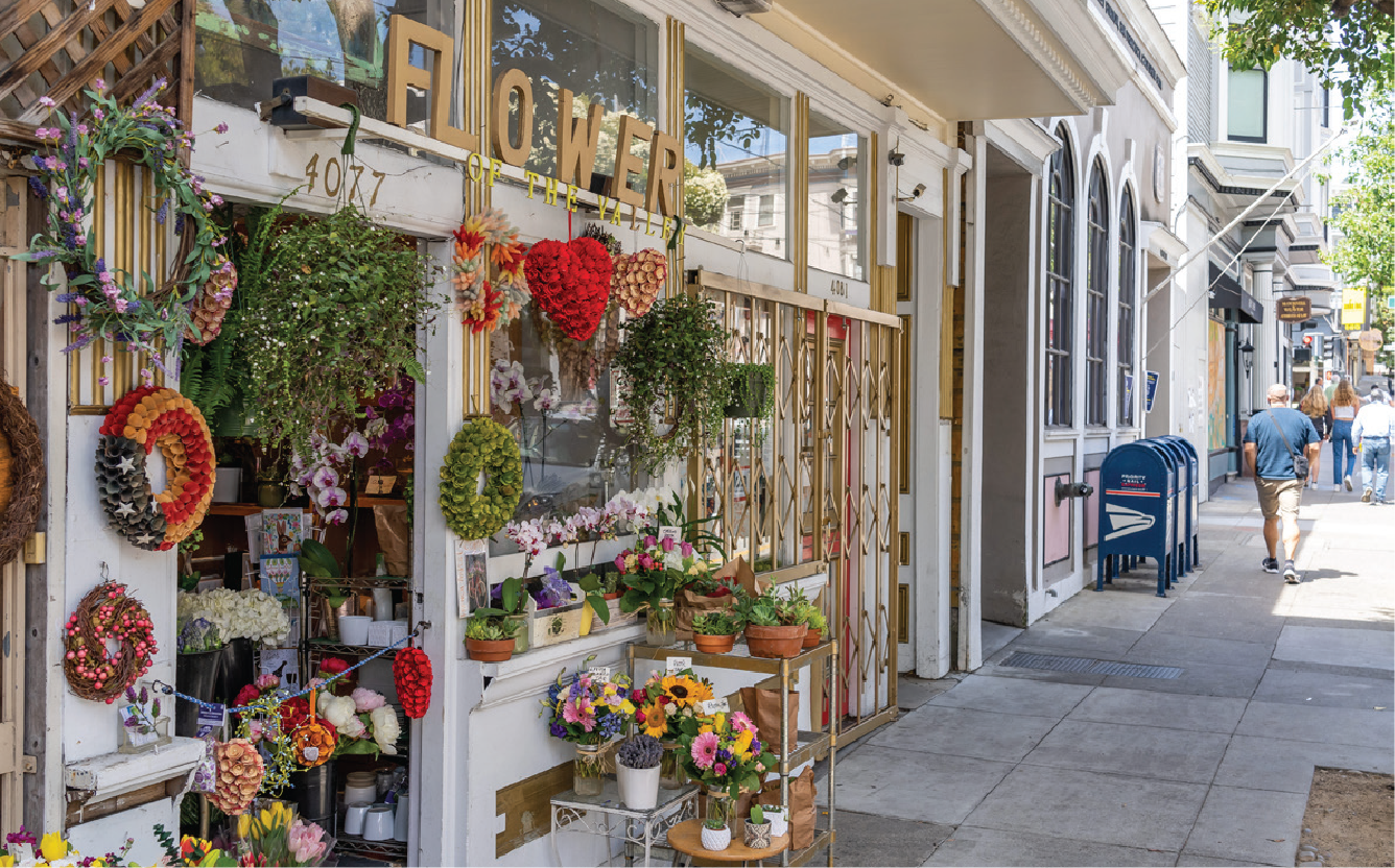 Flower shop storefront decorated with various floral arrangements and wreaths, with golden 'FLOWER' sign above the window, on a city sidewalk with pedestrians.
