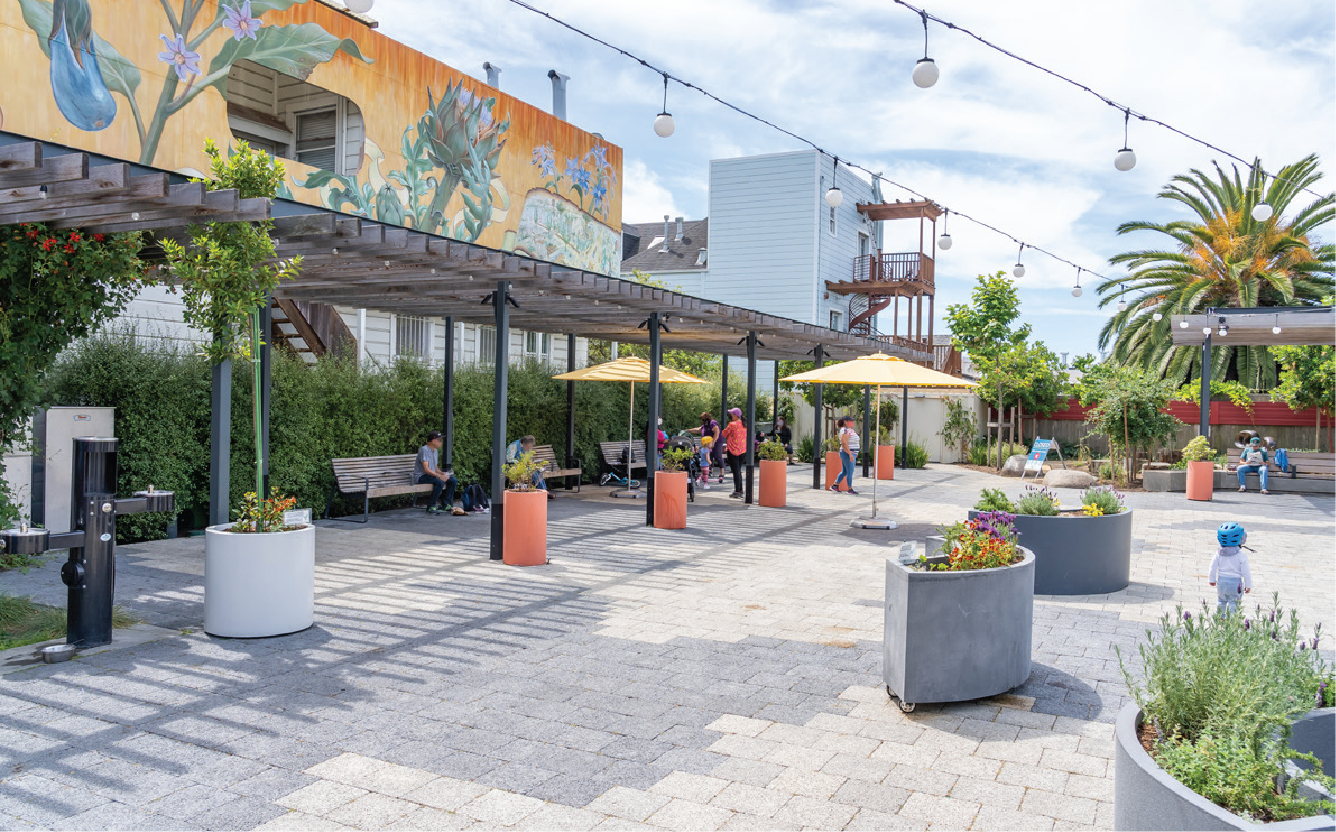 People sitting and walking in an outdoor urban park with benches, umbrellas, large planters, palm trees, and overhead string lights, with a mural painting on a building in the background.