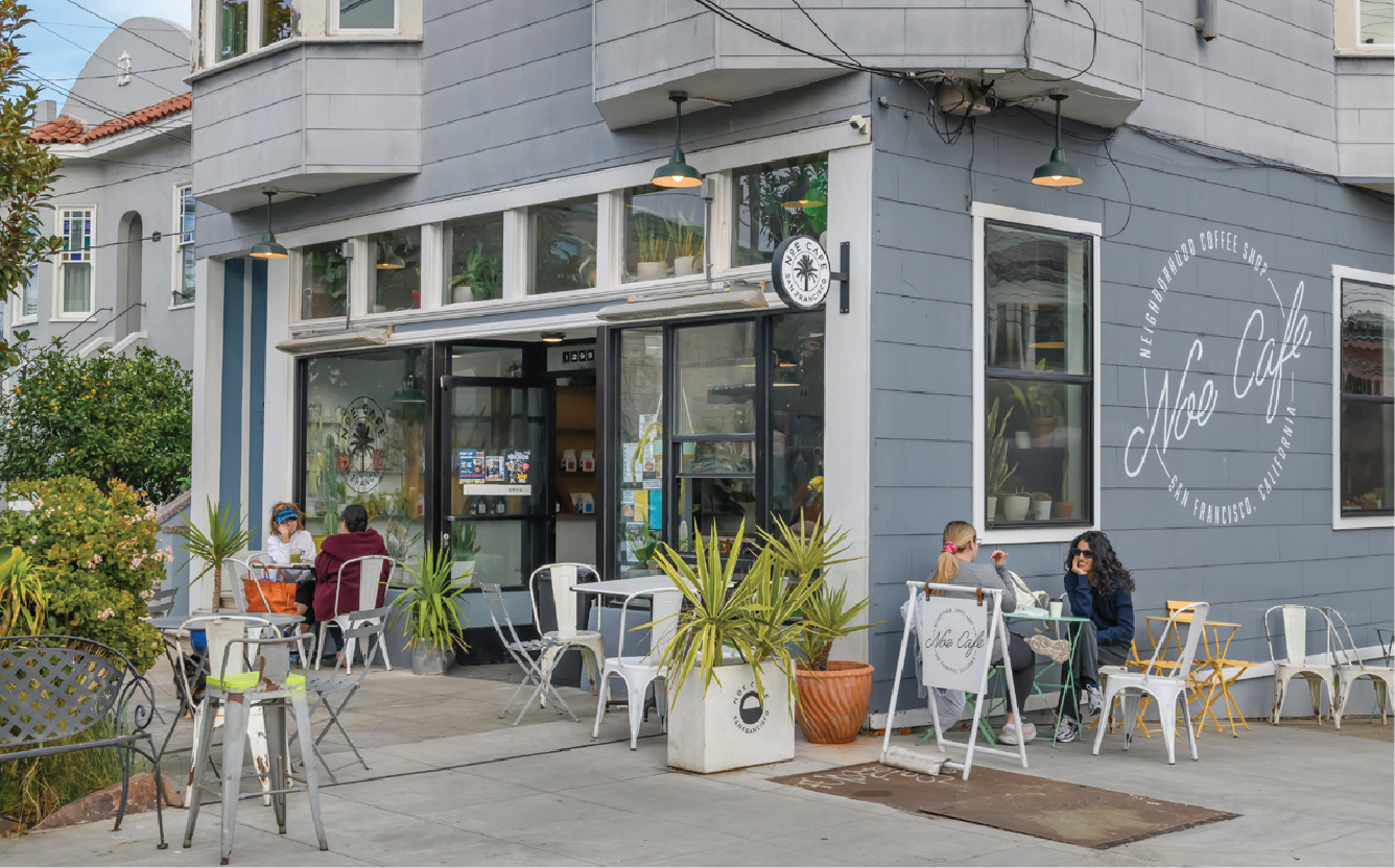 Exterior of Moe Cafe with outdoor seating, potted plants, and people sitting at tables in front of a gray building with large windows and a sign reading Moe Cafe, a neighborhood coffee shop in San Francisco, California.
