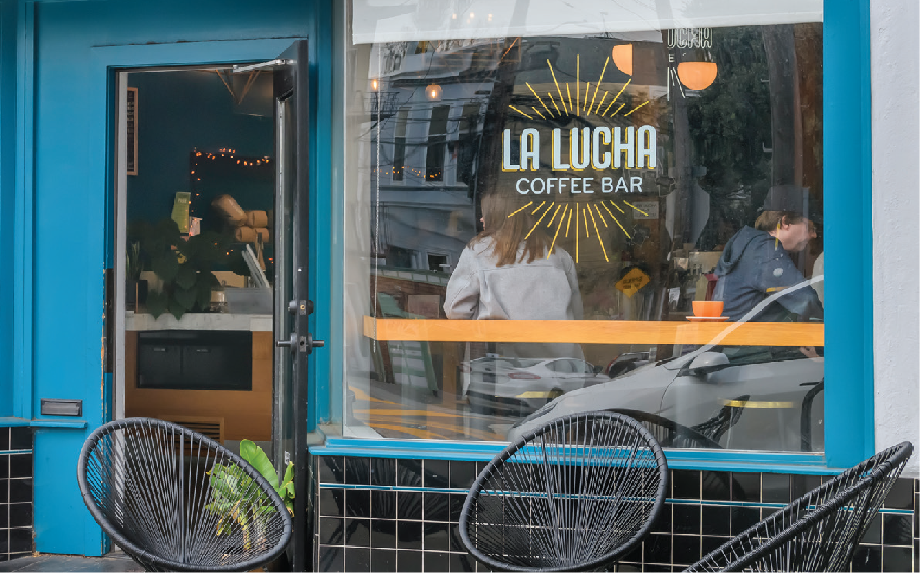 Front view of La Lucha Coffee Bar with outdoor seating, blue exterior, and large window featuring the logo and two people inside at the counter.