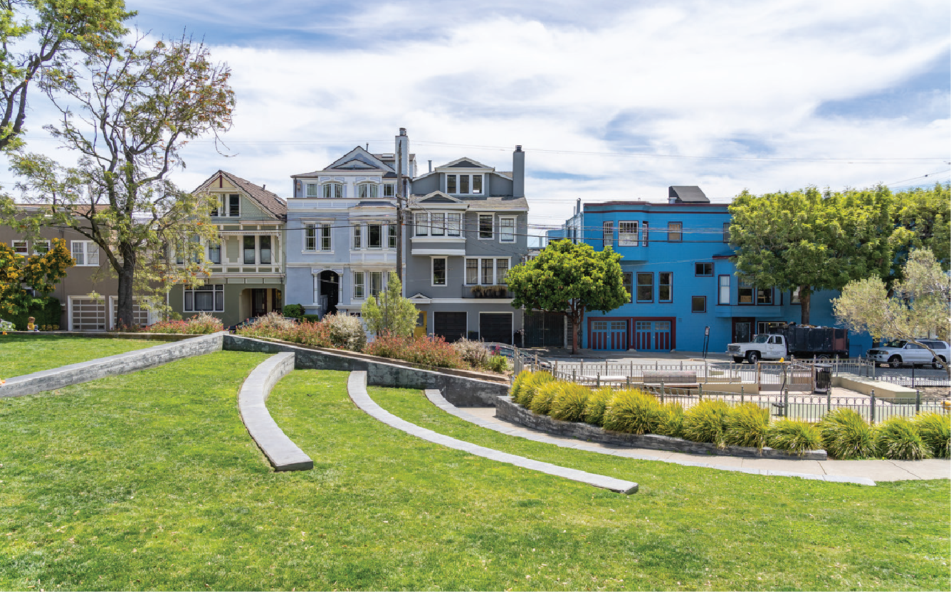 A park with curved concrete benches and green grass, white and gray houses with multiple stories and gardens in the background, trees, and a partly cloudy sky.