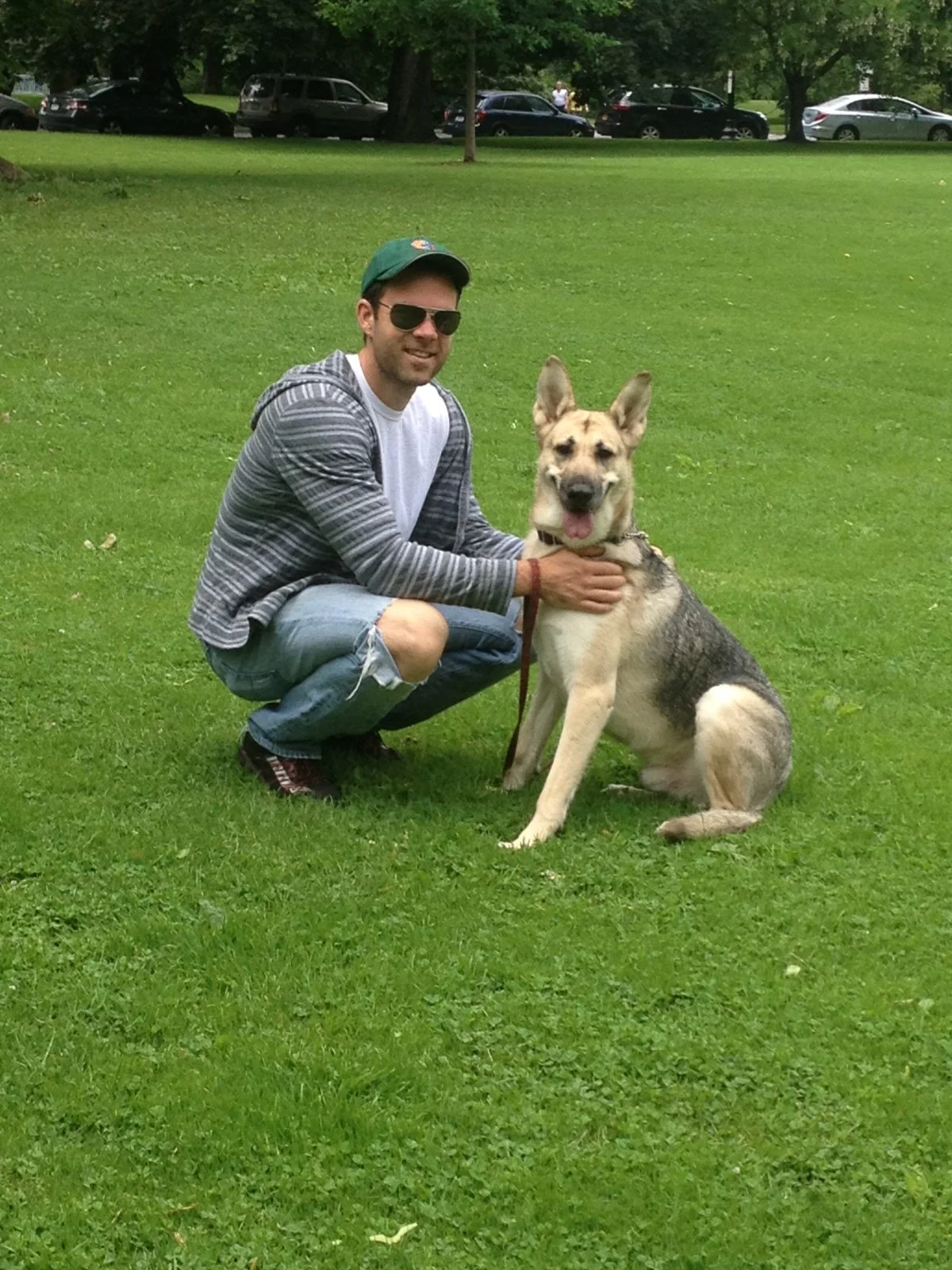 A man wearing sunglasses, a green cap, and a striped hoodie crouches next to a sitting German Shepherd dog on a grassy field in a park.