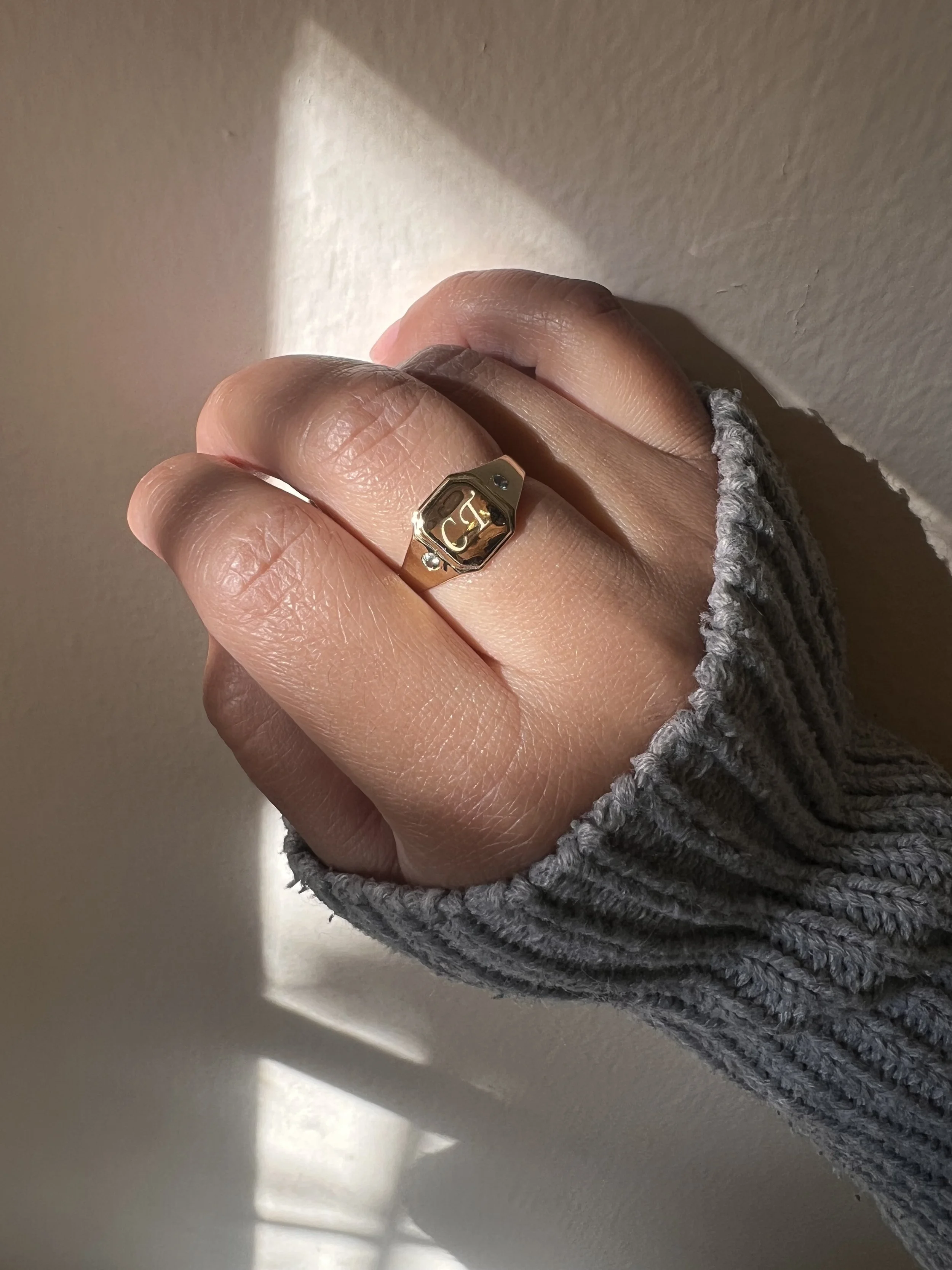 Close-up of a hand wearing a gray knitted sleeve, showing a gold ring with a design, resting against a light-colored wall with sunlight and shadows.