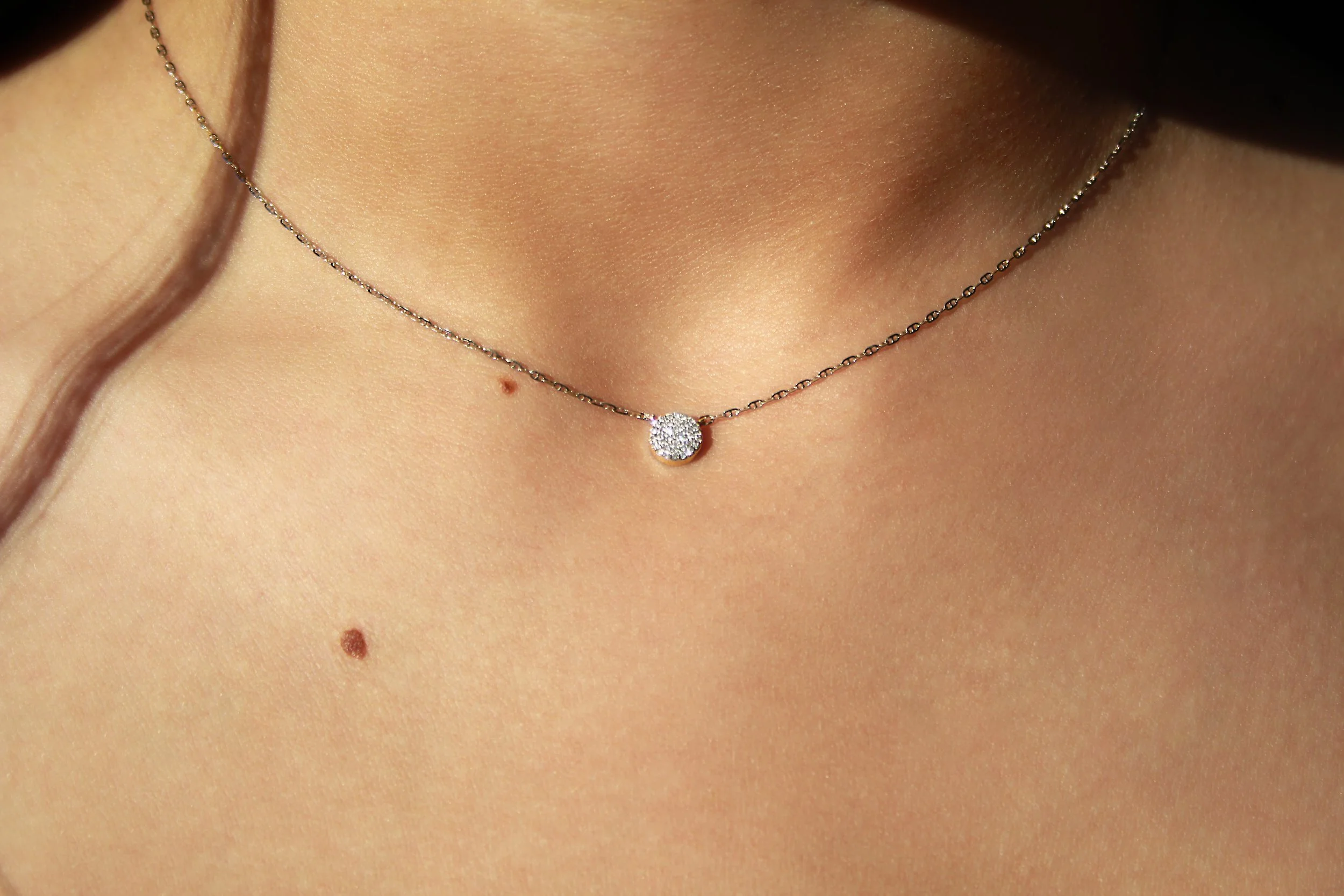 Close-up of a delicate silver necklace with a small, round, diamond-encrusted pendant on a person's neck, showing skin and a mole.