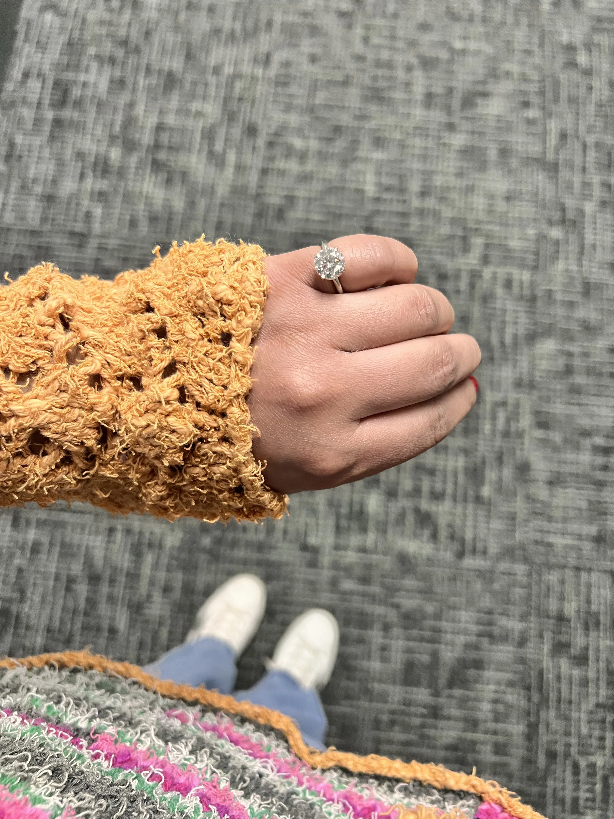 A person's hand wearing an engagement ring with a large diamond, they are dressed in a mustard-colored, textured jacket, and are standing on a gray carpeted floor. The photo is taken from a top-down perspective, showing the person's shoes, a colorful