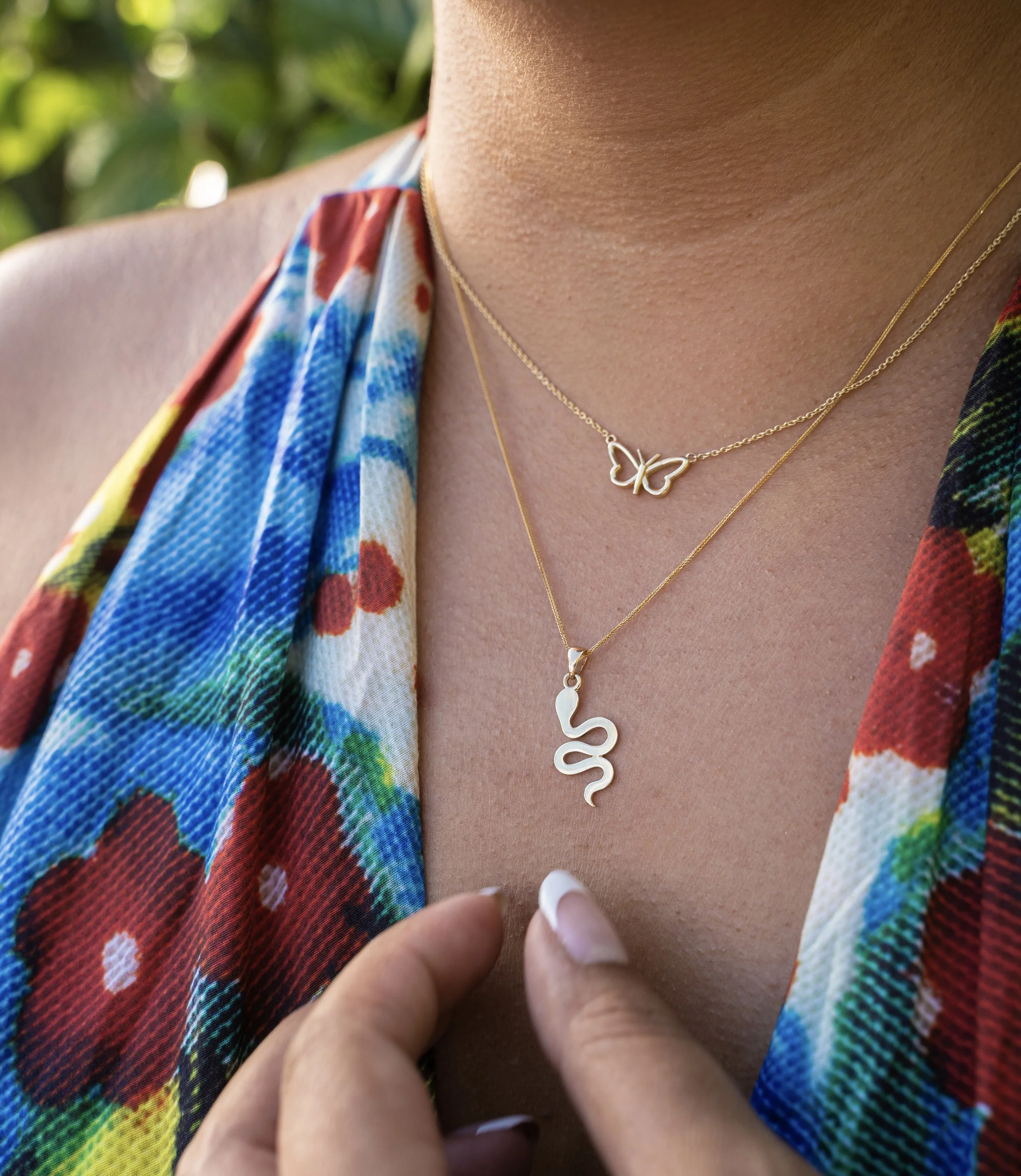 Close-up of a woman's neck wearing two layered gold necklaces, one with a butterfly pendant and the other with a snake pendant, with a colorful floral dress.