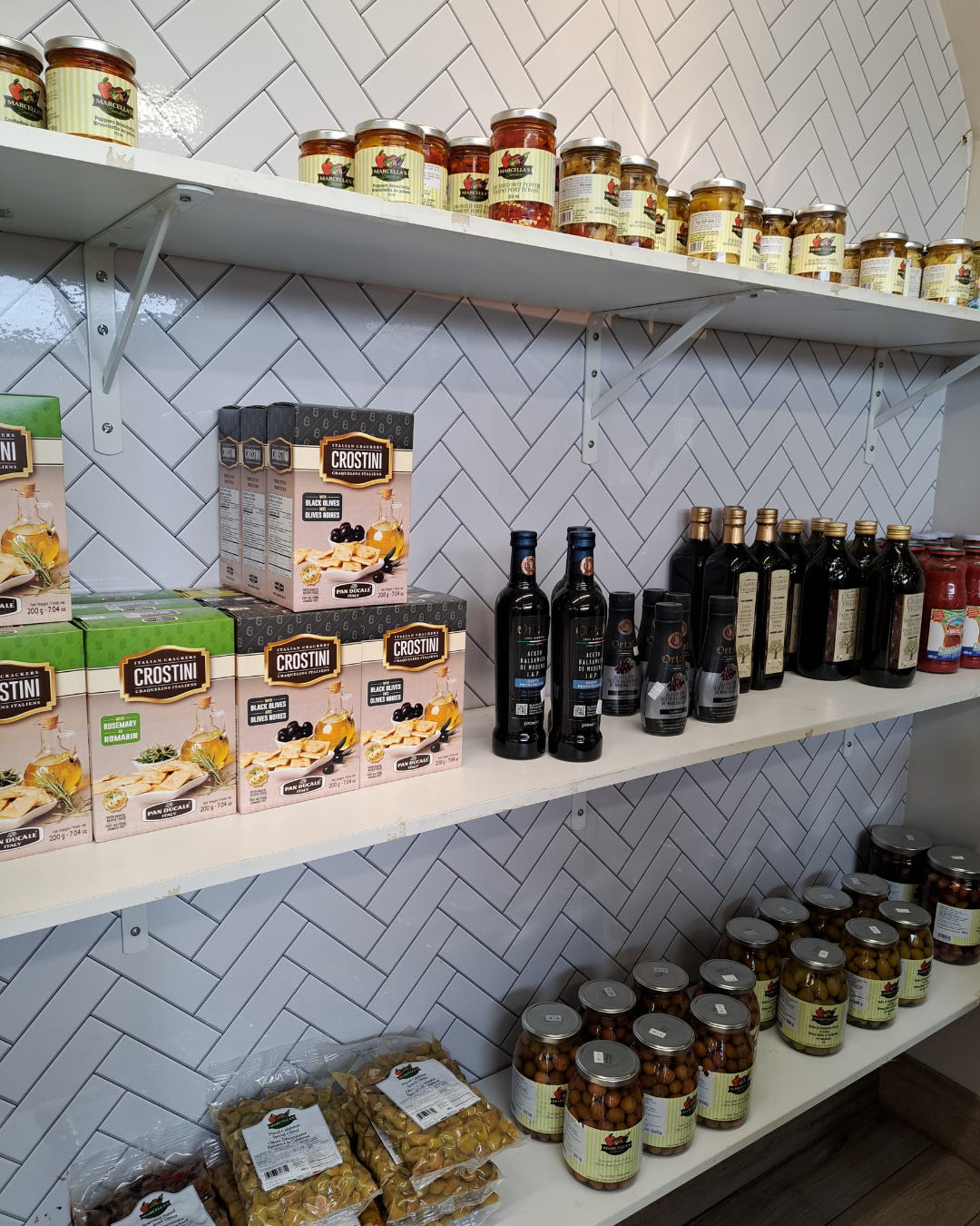 Shelves stocked with jars of olives, bottles of oil, and packaged crackers against a white tiled wall.