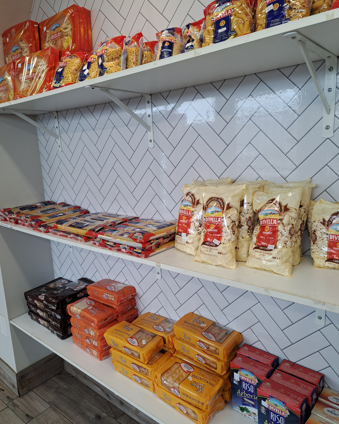 Shelves stocked with packages of pasta and rice in a store with white herringbone tile background.