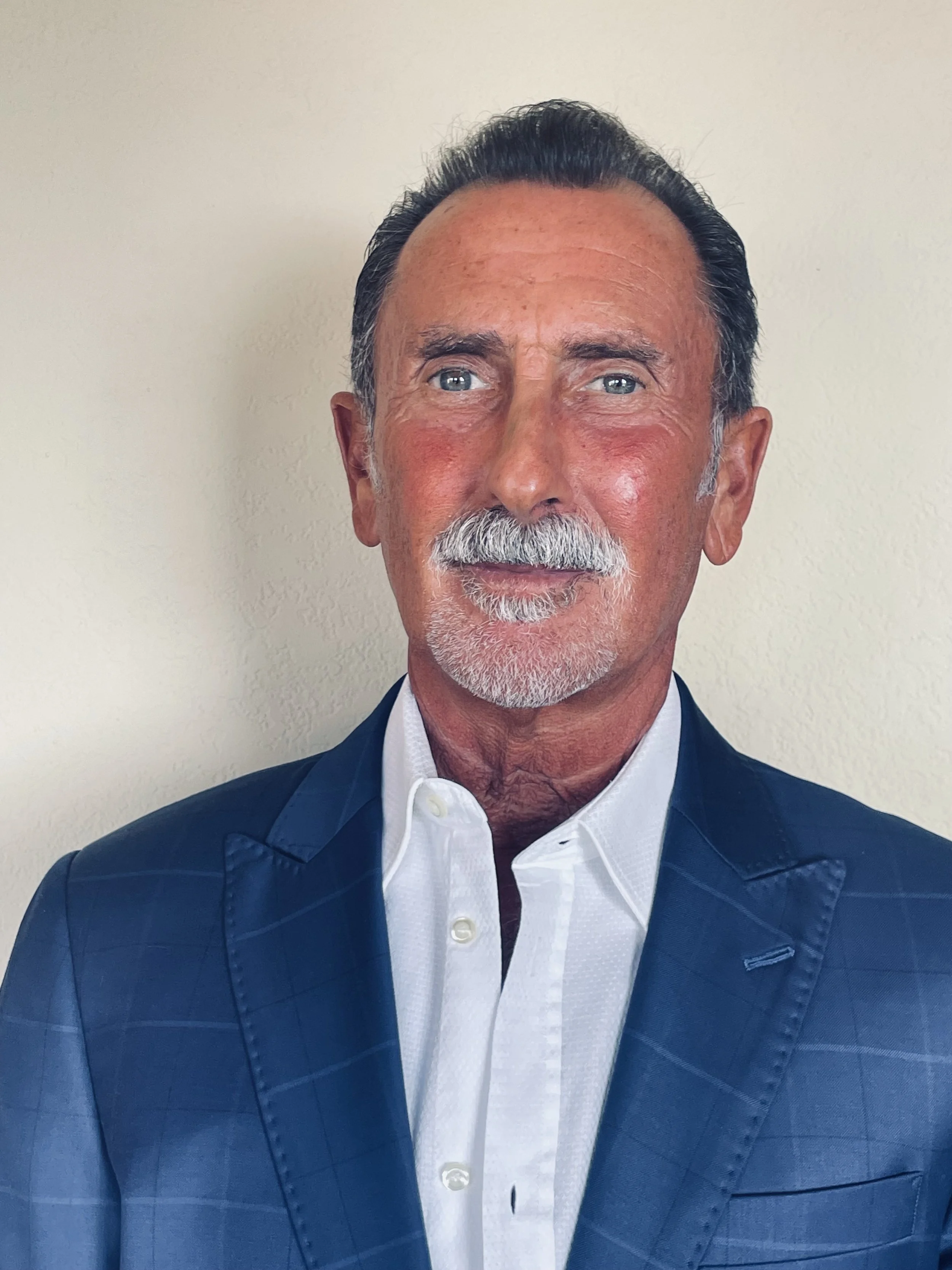 A portrait of a mature man with blue eyes, gray hair, and a gray mustache. He is wearing a dark blue suit jacket and a white dress shirt, standing against a plain light-colored background.