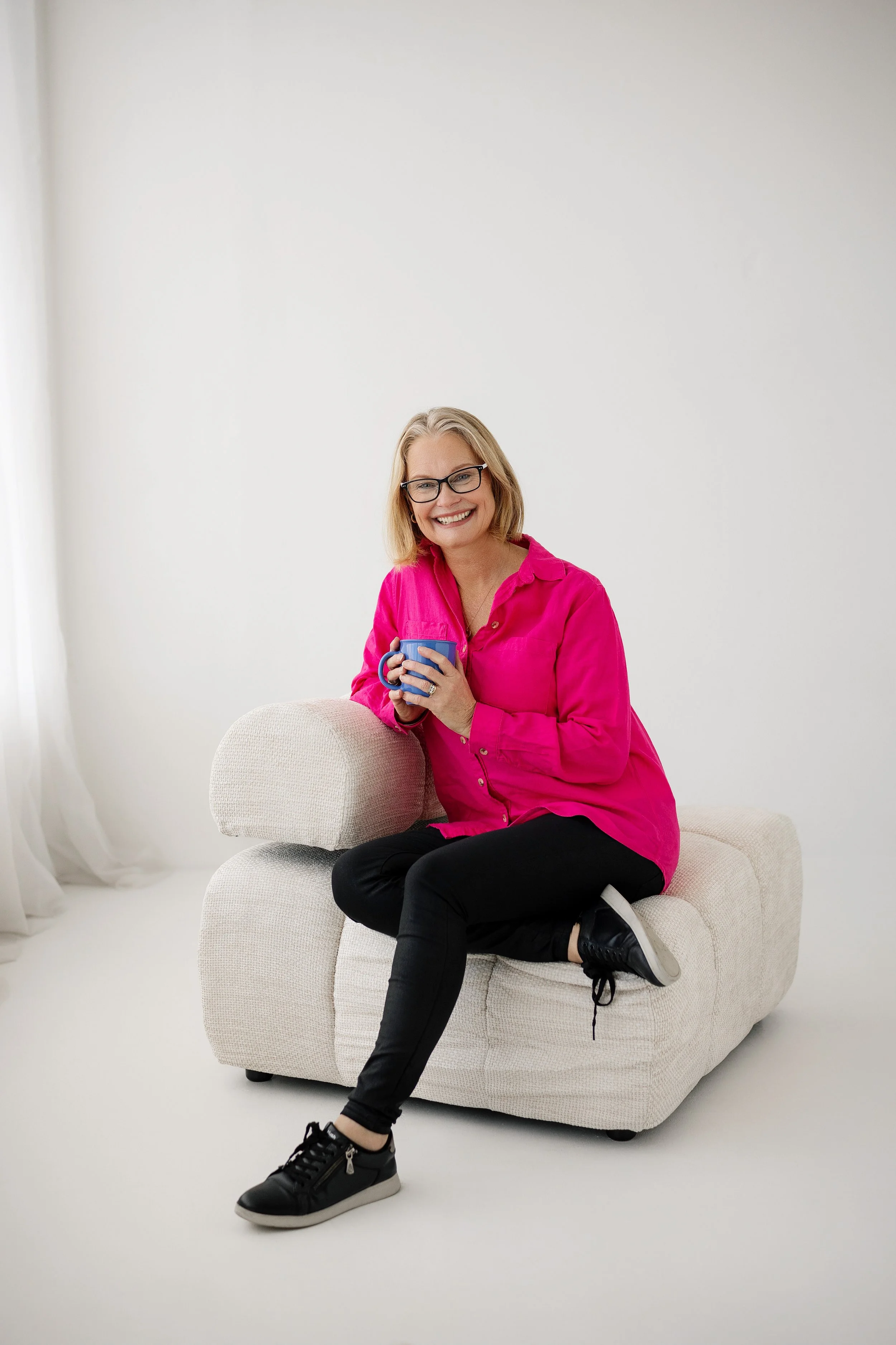 Vicki, a friendly, cheerful woman wearing a hot pink button up shirt and black pants sitting comfortably in a soft chair holding a bright blue mug of tea smiling welcomely