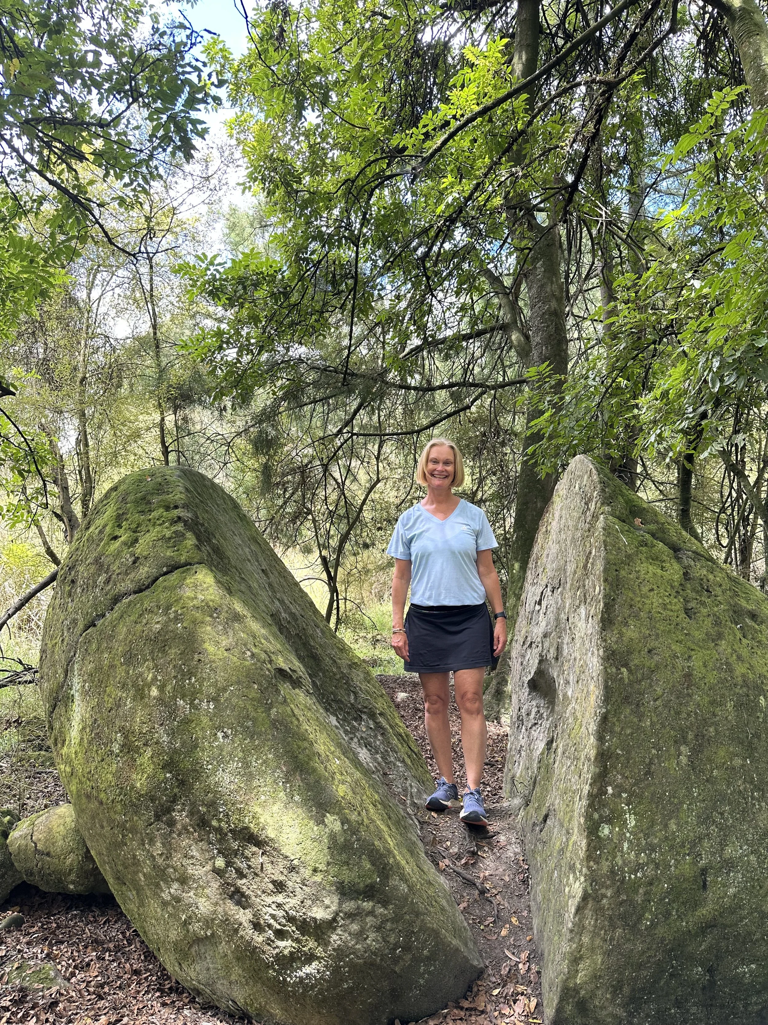 A woman, Vicki walking in beautiful soothing nature by a huge rock that has been split in half and is covered in moss and lichen. Come as you are, leave feeling lighter, brighter, and more energised.