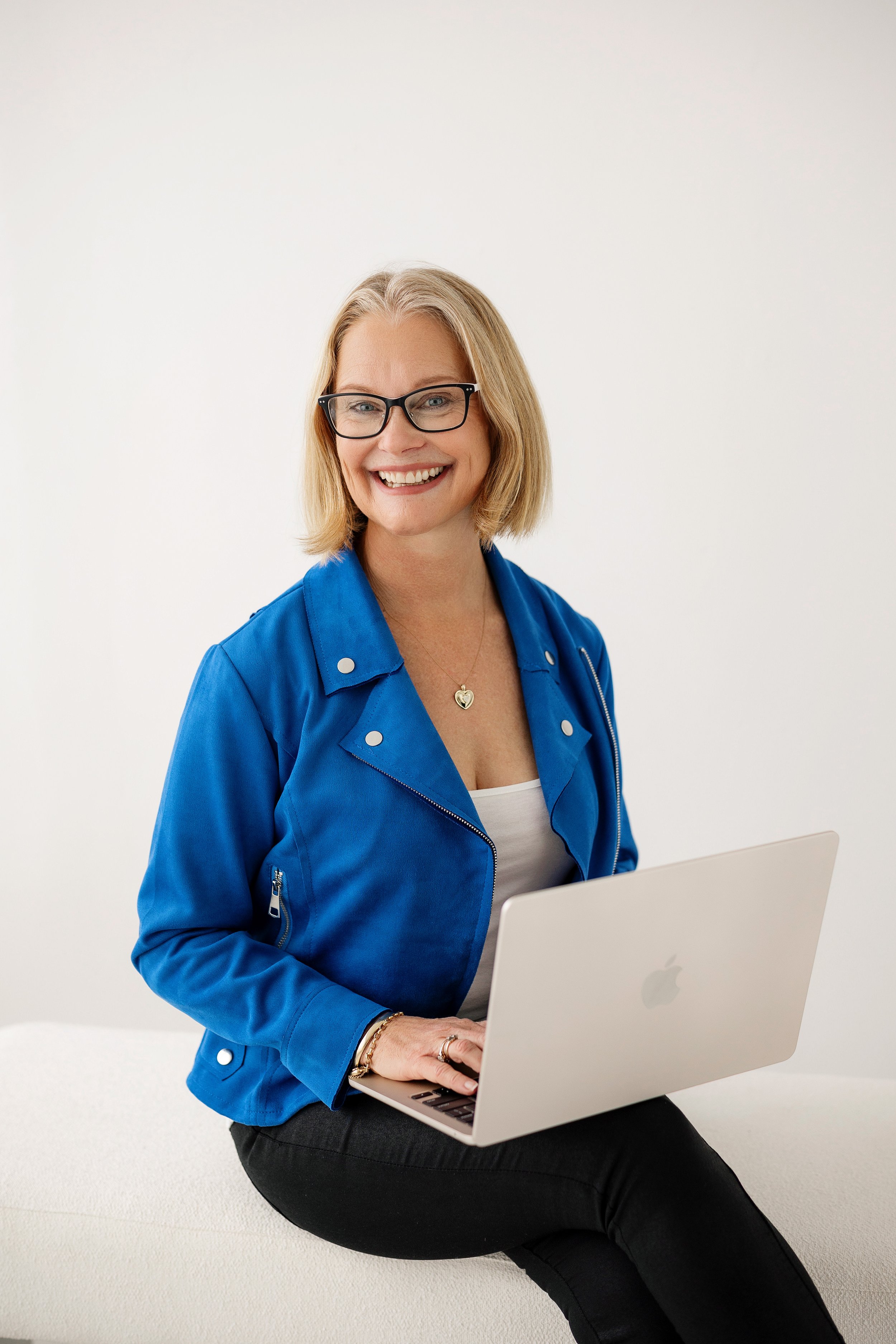 Vicki - a woman with blonde hair wearing glasses, a blue jacket, and a white top, sitting on a white surface, holding a silver MacBook laptop, and smiling, so happy in her job as a Health Coach.