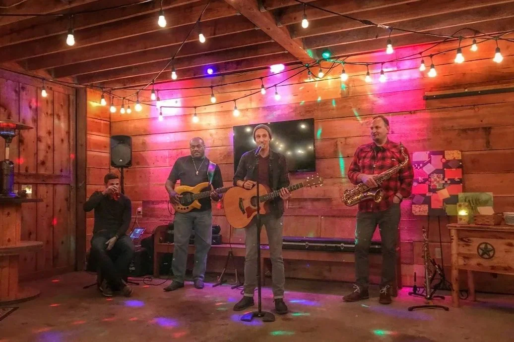 Four men performing live music on a small indoor stage with wooden walls and colorful string lights, including a guitarist, bassist, saxophonist, and a singer with an acoustic guitar.