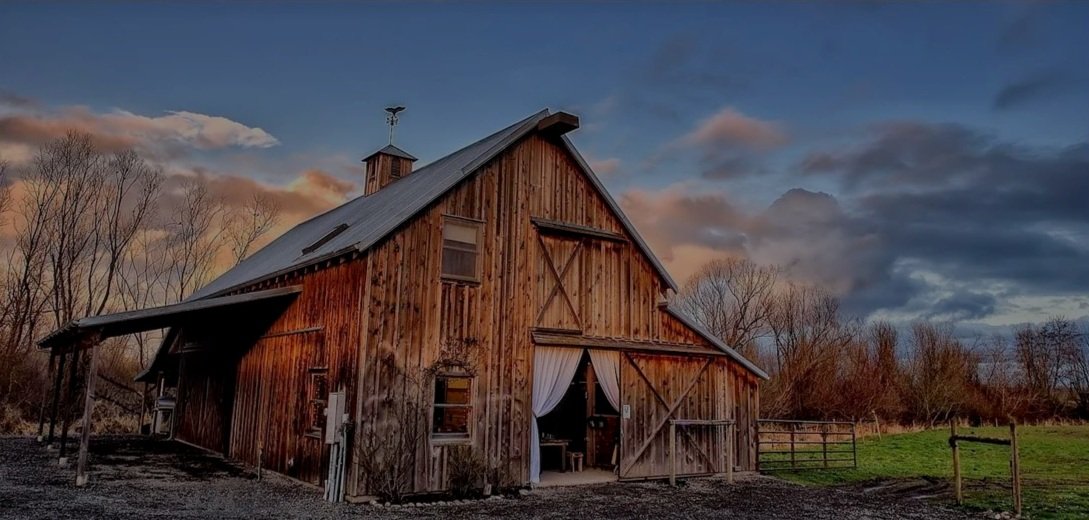 A rustic wooden barn with a weather vane on top, set against a partly cloudy evening sky, surrounded by trees and open fields.