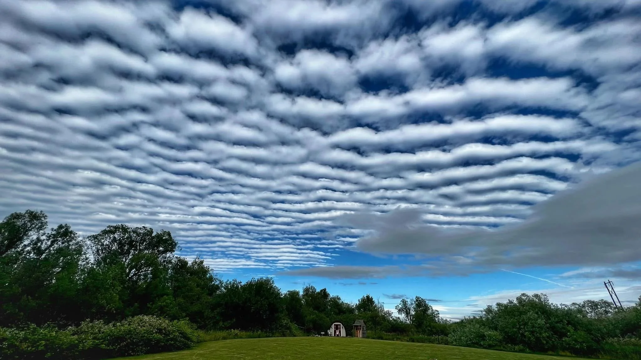 Cloudy sky with parallel banded clouds over a green landscape with trees and small buildings.