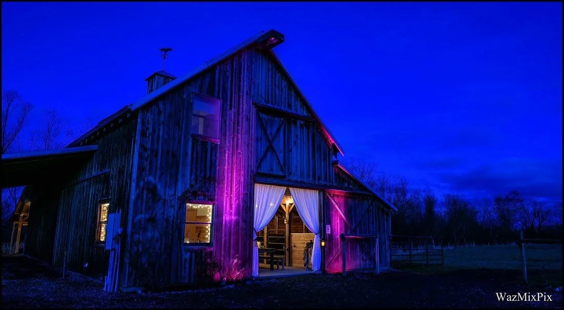 A rustic wooden barn with colorful purple and pink lighting at dusk, with open doors revealing an interior decorated for an event.