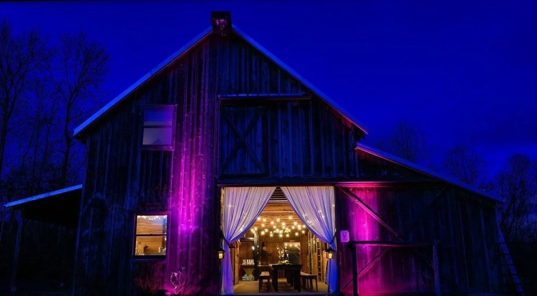 Barn with open doors, curtains, string lights inside, and purple and pink lighting outside at night.
