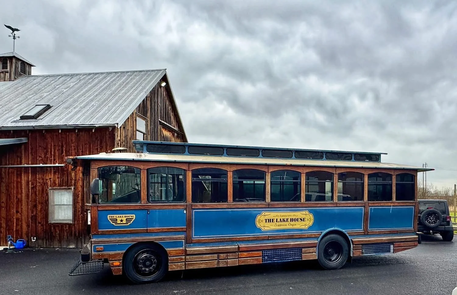 A vintage blue and wooden trolley bus with signage says 'The Lake House, Champagne Oregon,' parked outside a rustic wooden building on a cloudy day.