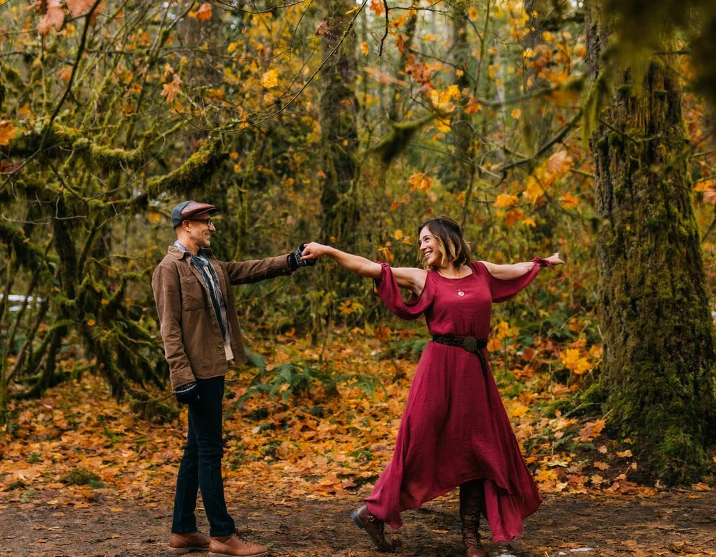 A man and a woman holding hands and dancing in a forest with autumn leaves.